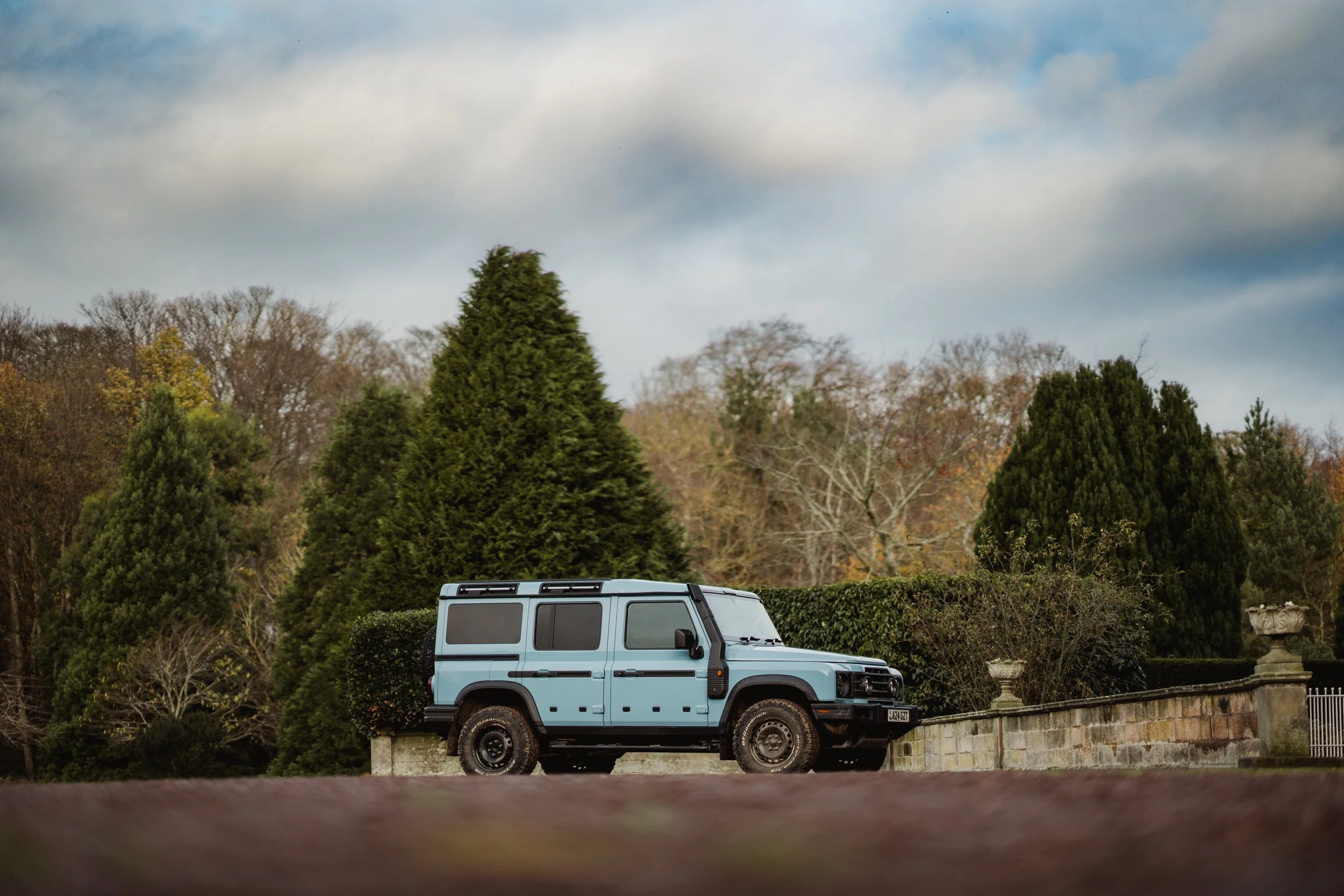 A blue Mercedes-Benz G-Class SUV parked on a stone ledge in front of a hedge, with tall trees and an overcast sky in the background.