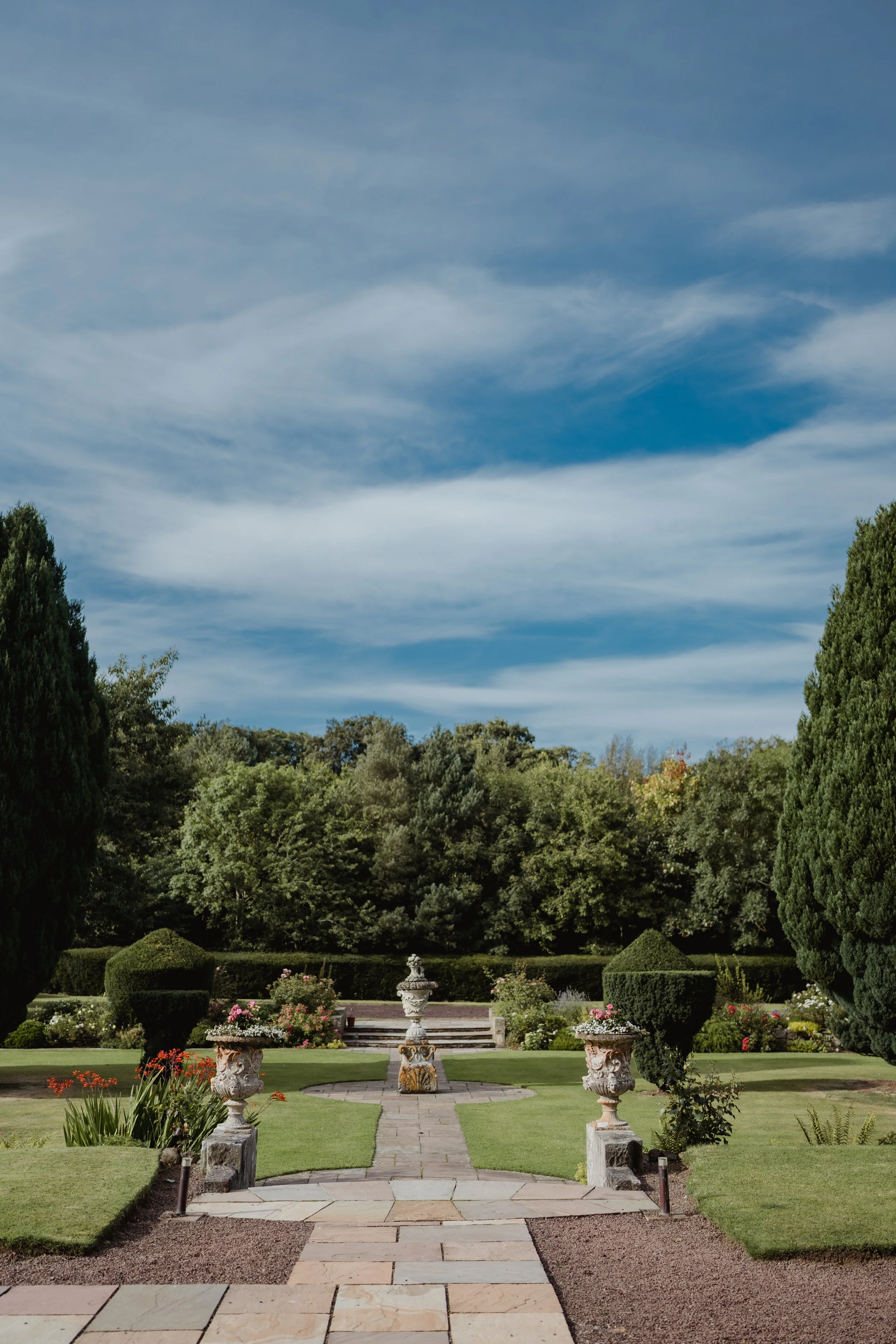 A formal garden with a stone pathway, decorative urns, green lawns, flower beds, and a fountain, surrounded by tall trees under a partly cloudy sky.
