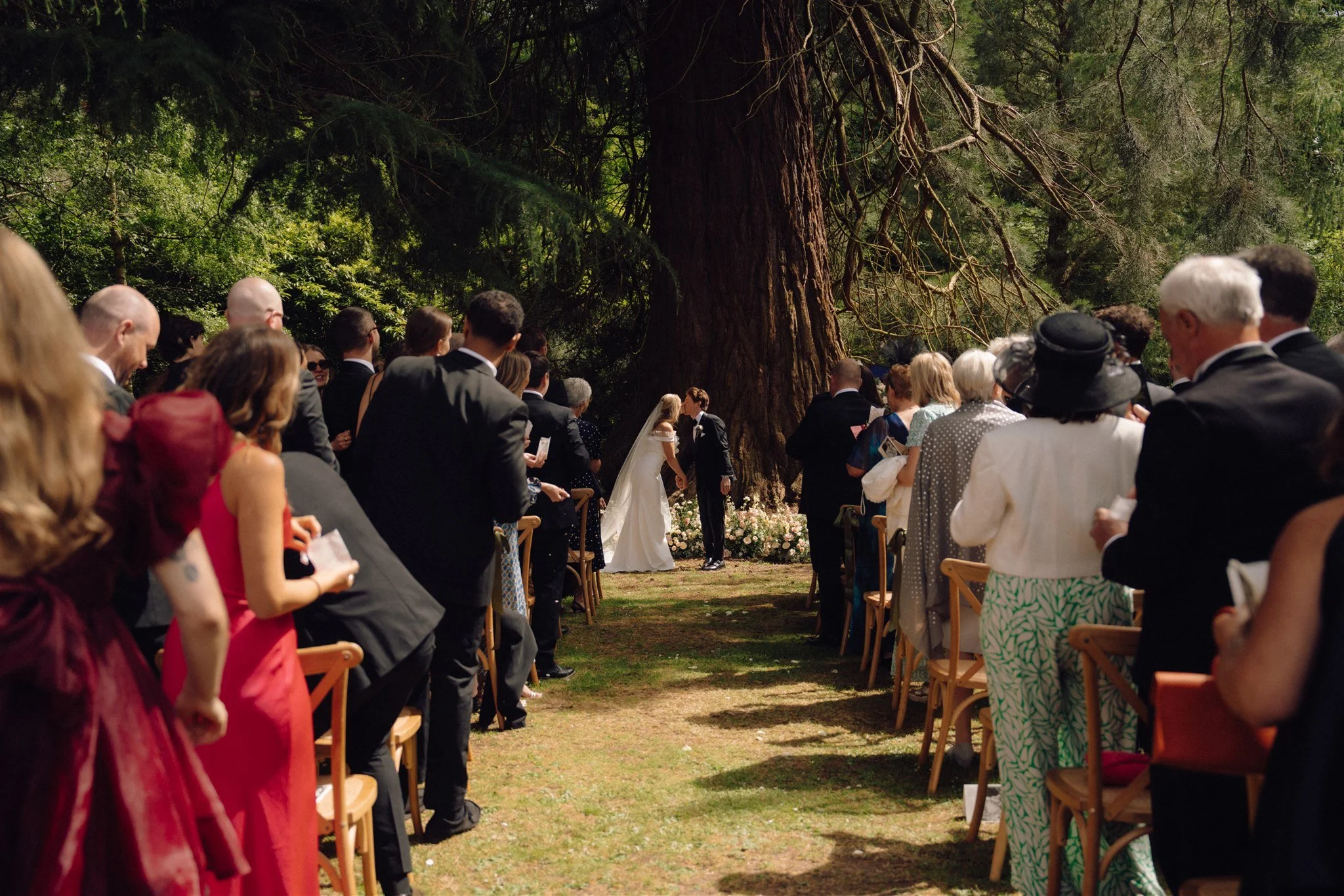 Outdoor wedding ceremony with guests seated on wooden chairs under large trees, with the bride and groom in the center exchanging vows.