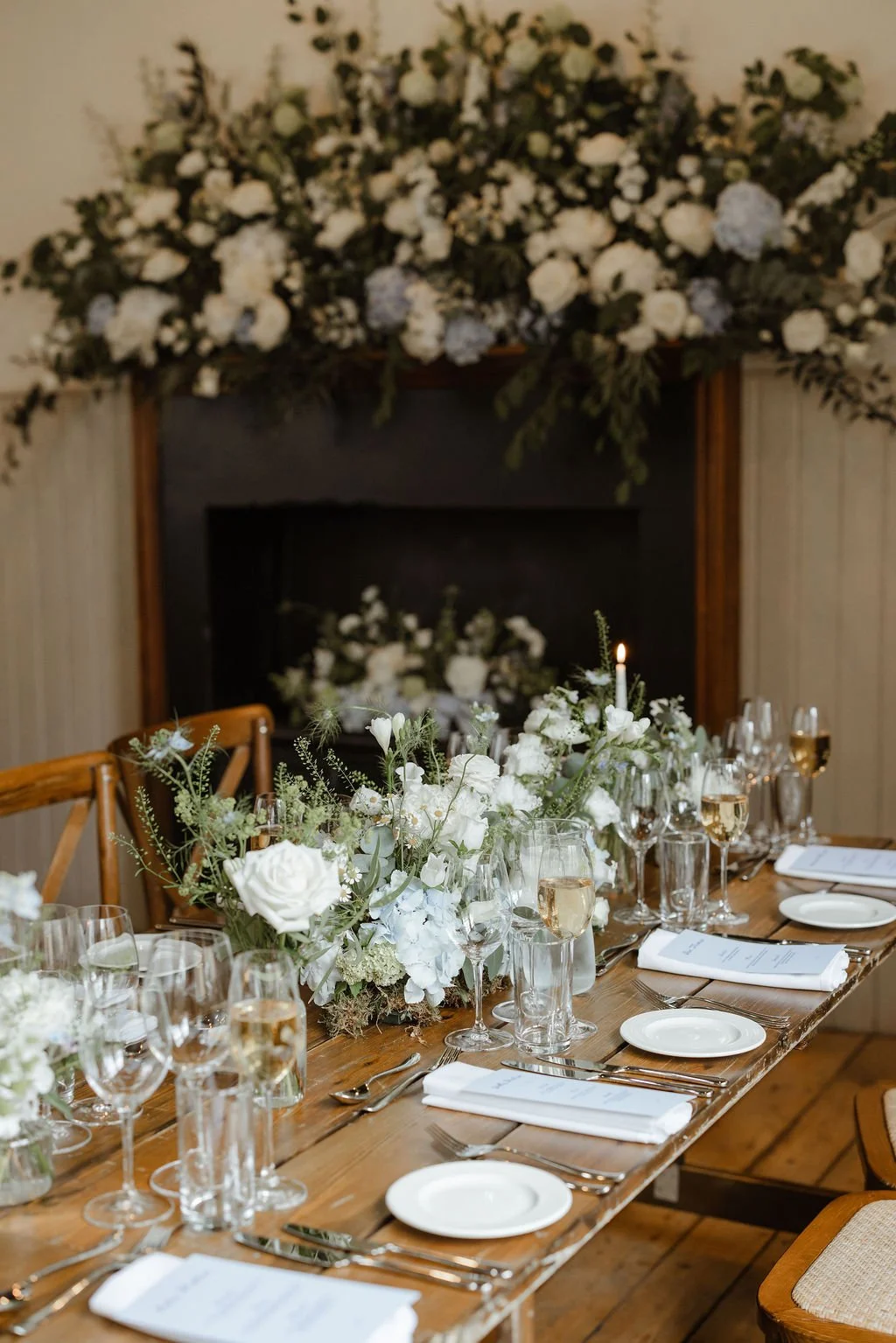 Elegant dining table decorated with white flowers, candles, and glassware for a wedding or special event, with a large floral arrangement on the mantel in the background.