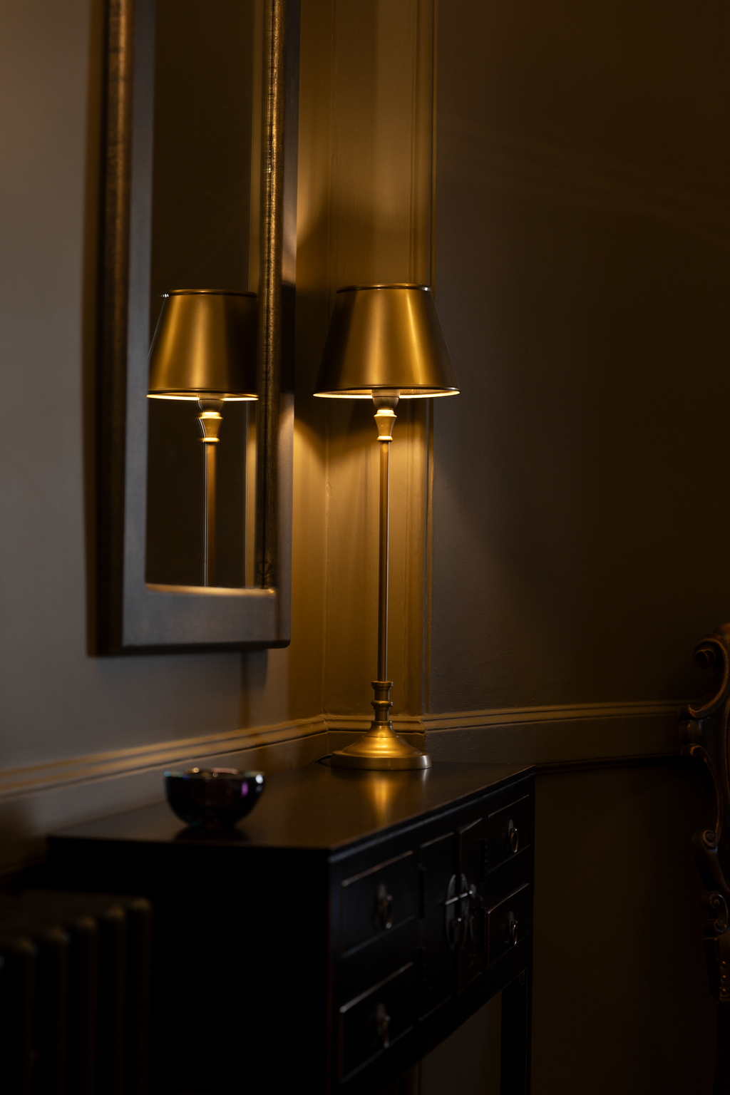 A gold table lamp with a matching gold lampshade standing on a black sideboard, reflected in a mirror on the wall in a dimly lit room.