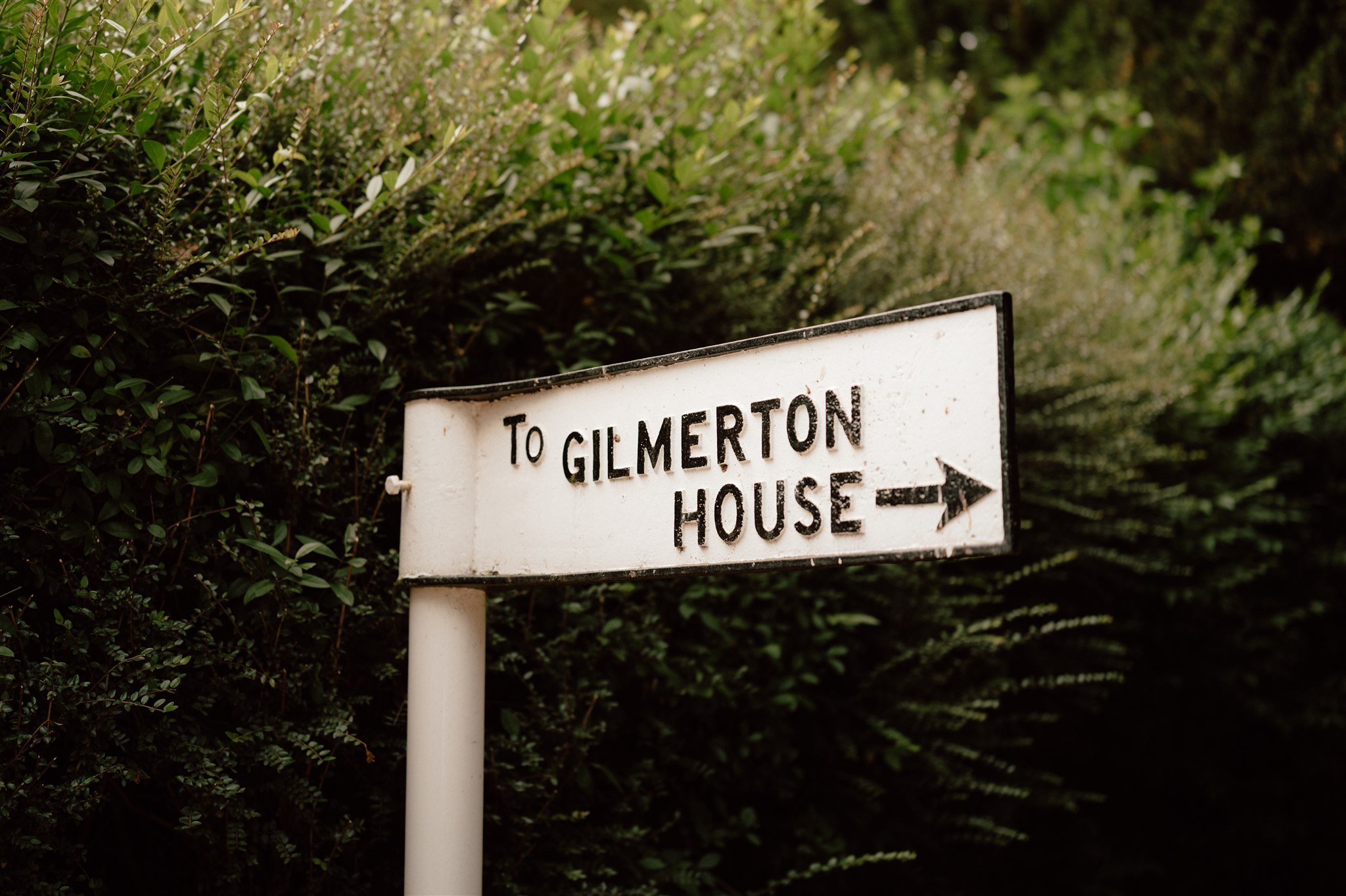 A white directional sign with black text and an arrow pointing right, indicating the way to Gilmerton House, surrounded by green foliage.