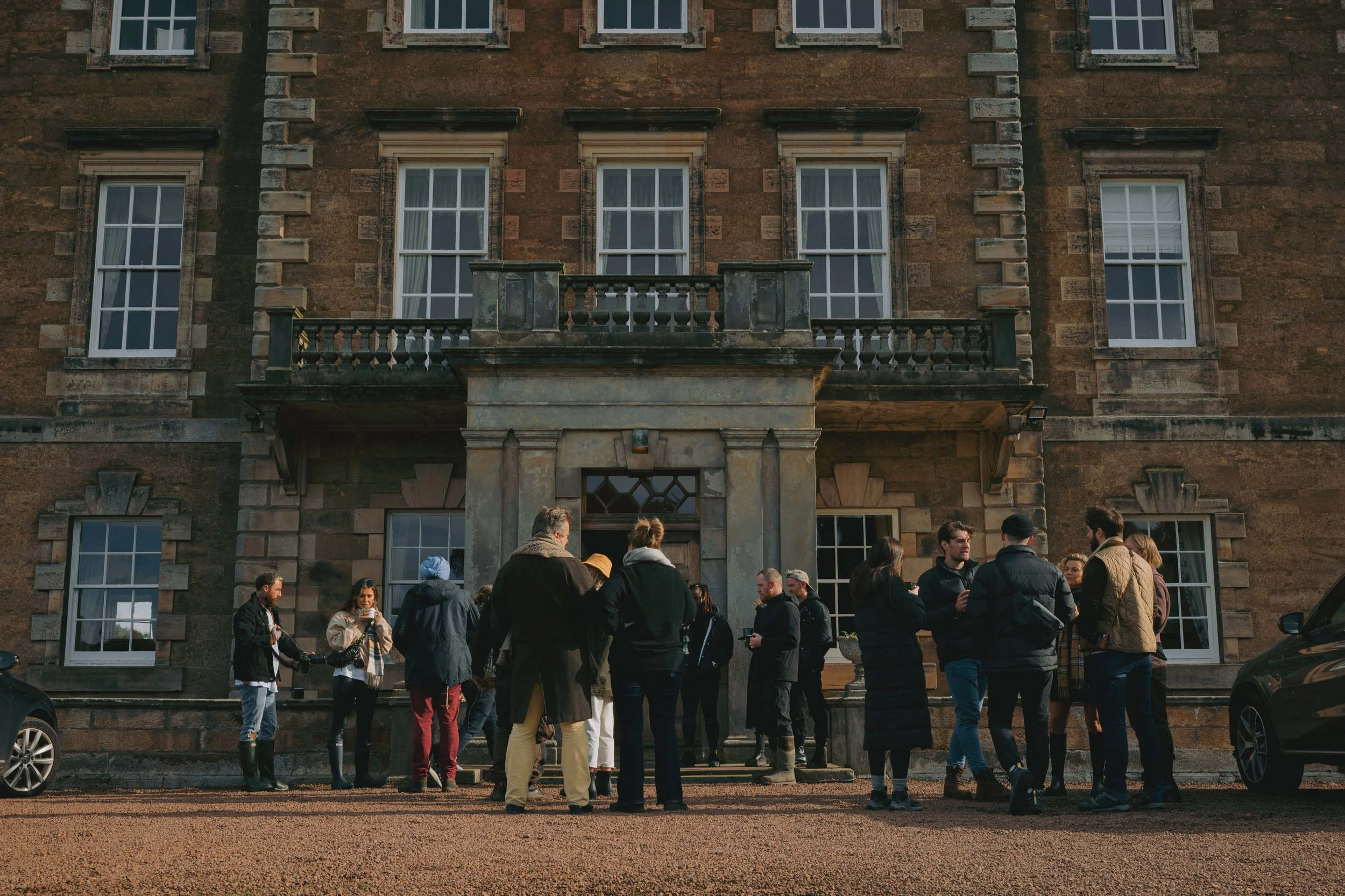 A group of people gathered outside a large, historic brick building with multiple windows and a stone balcony. Some are talking and holding drinks. The scene appears to be social or a casual gathering.