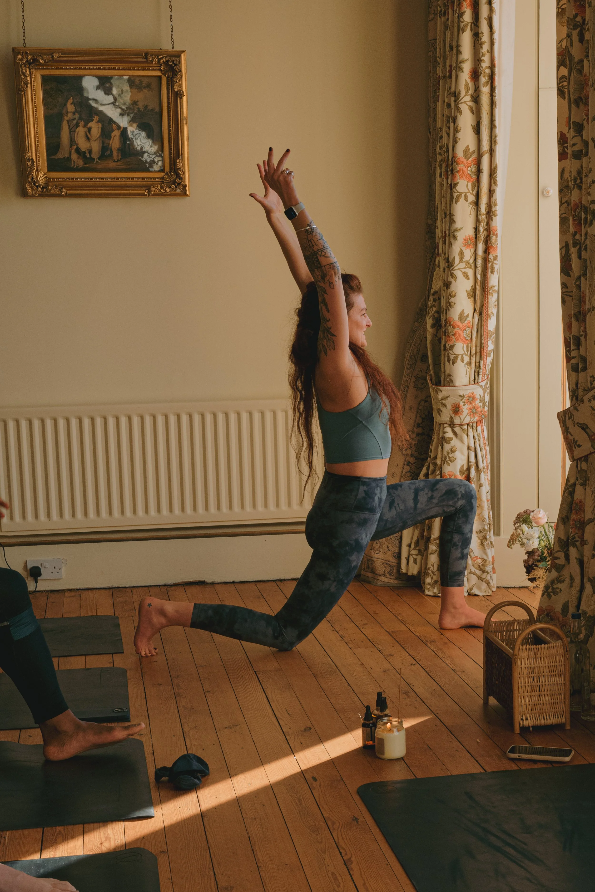 A woman practicing yoga indoors, kneeling on one knee with the other leg bent, one arm reaching upwards, and the other resting on her knee. She has long, wavy hair and tattoos on her arm, and is wearing a sports bra and patterned leggings. The room has wooden flooring, floral curtains, a framed painting on the wall, and yoga mats on the floor.