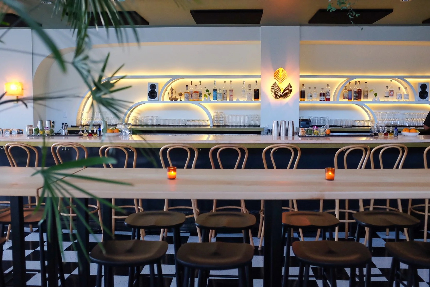 Wide shot of a sculptural, back-lit, Art Deco inspired back bar and long pink marble bar top. There is a long, communal table made of blonde wood is in the foreground.