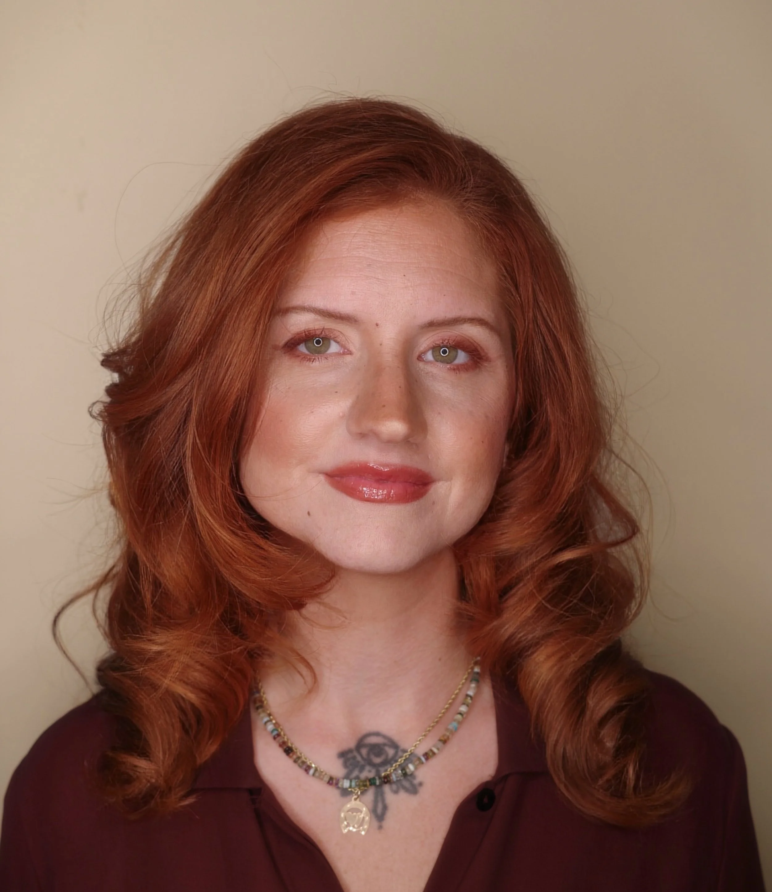 Close-up of a woman's face and upper body with red curly hair, wearing a dark top and layered necklaces, standing in front of a plain beige wall.