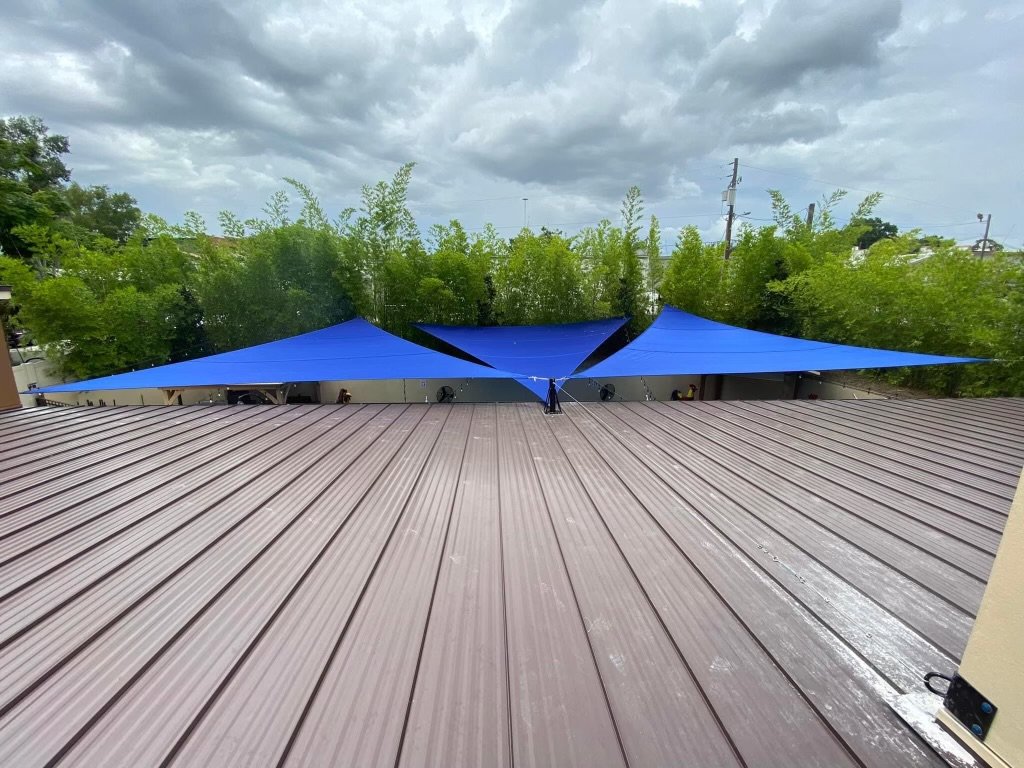 Overhead view of vibrant blue triangular shade sails installed above an outdoor area, providing extensive sun coverage and a modern aesthetic. Set against a backdrop of lush green bamboo, the shade sails create a striking contrast, offering both func