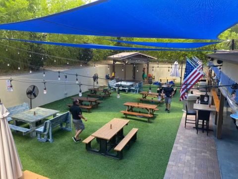 Inviting outdoor dining space covered with vibrant blue shade sails, providing ample sun protection over picnic tables arranged on artificial turf. String lights hang across the area, creating a warm and festive ambiance. The setup includes American 