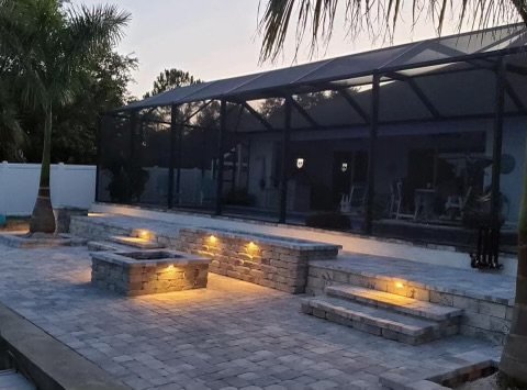 Beautiful custom hardscape design featuring multi-level stone pavers with built-in, low-voltage LED lighting, creating a warm ambiance at dusk. The outdoor living area includes a cozy, elevated fire pit space surrounded by lush tropical landscaping a