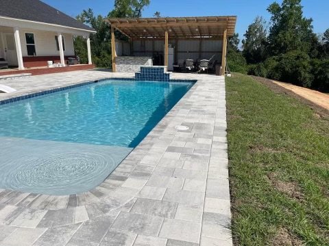 Beautiful backyard pool area with a custom paver deck, featuring a built-in sun shelf for relaxation and a waterfall feature for added elegance. Adjacent to the pool is a spacious pergola with an outdoor seating area and kitchen, ideal for entertaini