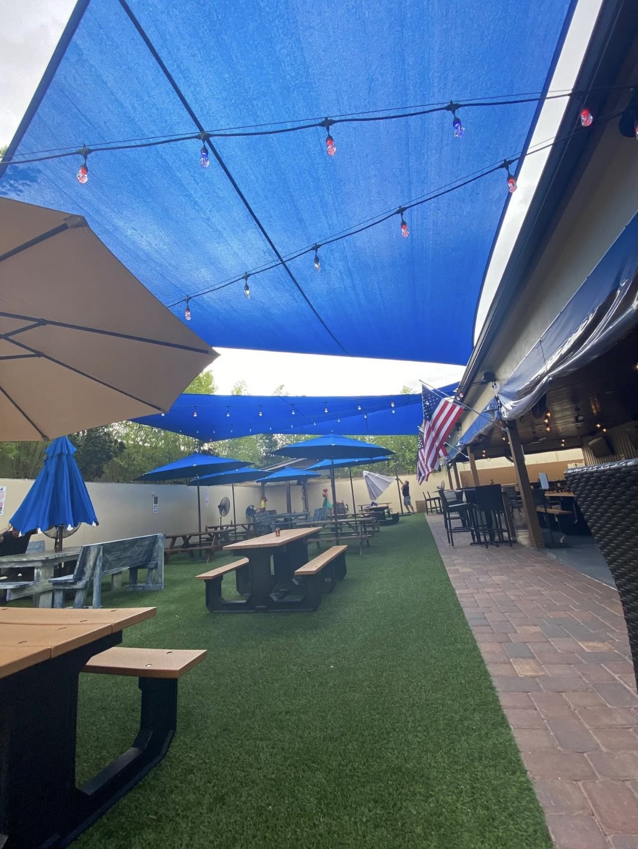 Vibrant outdoor seating area with large blue shade sails providing extensive sun protection over picnic tables and artificial turf. Festive string lights hang beneath the shade sails, adding a warm ambiance to the space, making it ideal for gathering
