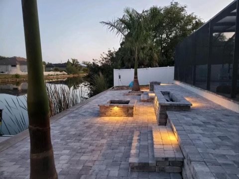 Modern lakeside patio featuring custom stone pavers and ambient low-voltage LED lighting, creating a warm and inviting atmosphere. The multi-level seating area includes a built-in fire pit, perfect for gatherings by the water. Surrounded by lush palm