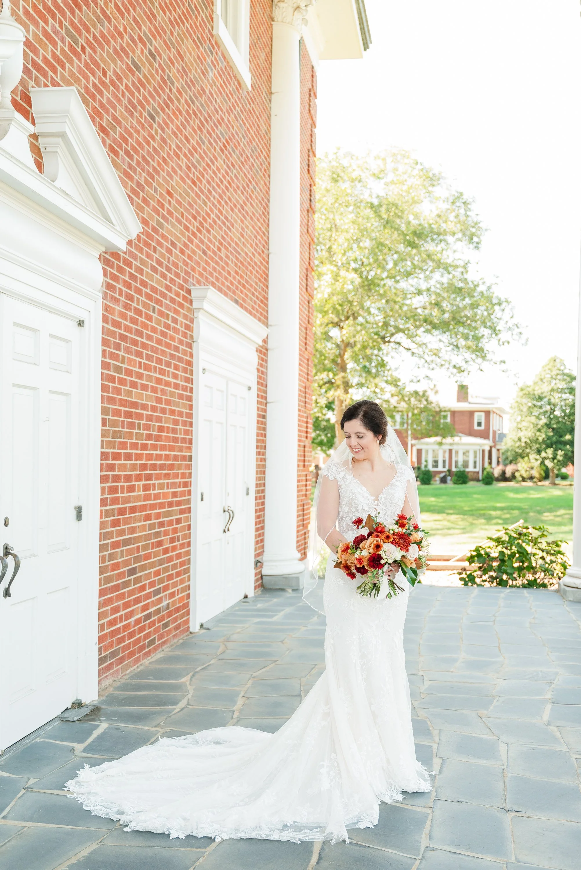 A bride in a white lace wedding dress holding a large bouquet of flowers, standing outside near a red brick building with white doors and classical architectural features, smiling and looking down.