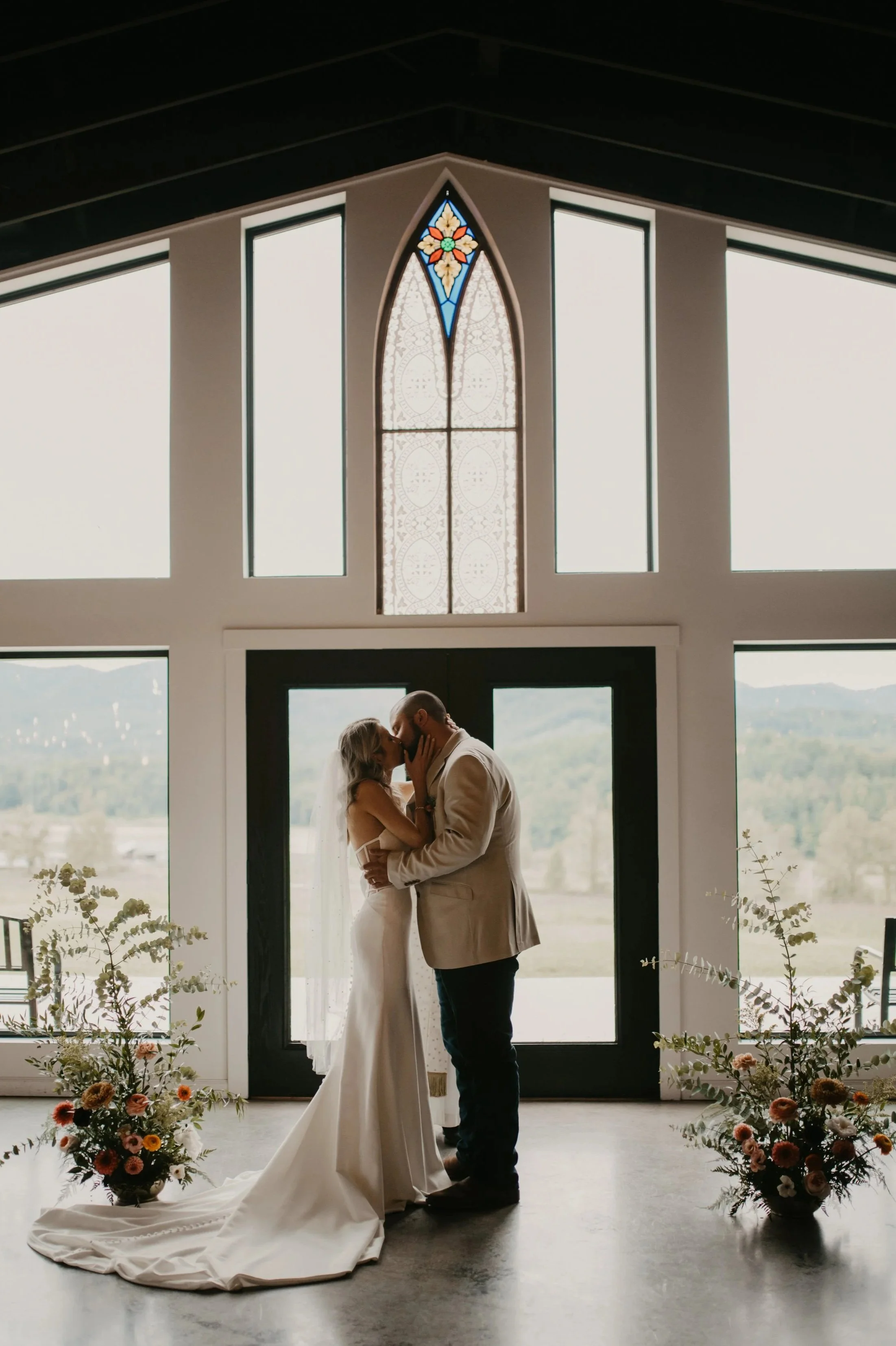 A couple sharing a kiss during their wedding ceremony inside a modern church, with large windows, floral arrangements, and stained glass window in the background.