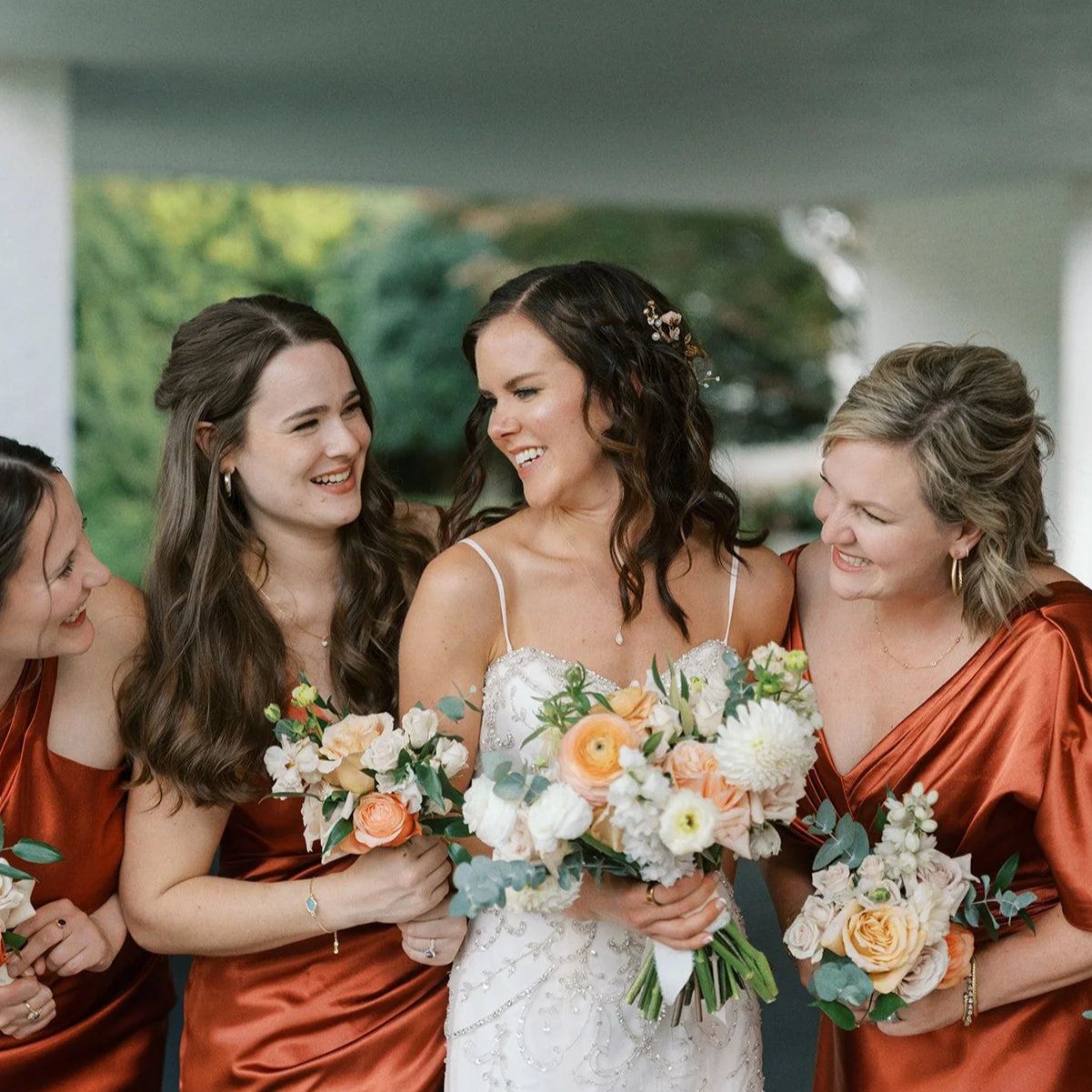 A bride with dark wavy hair in a white wedding gown holding a bouquet, smiling at her bridesmaids. The bridesmaids in rust-colored dresses are holding bouquets, laughing and smiling.
