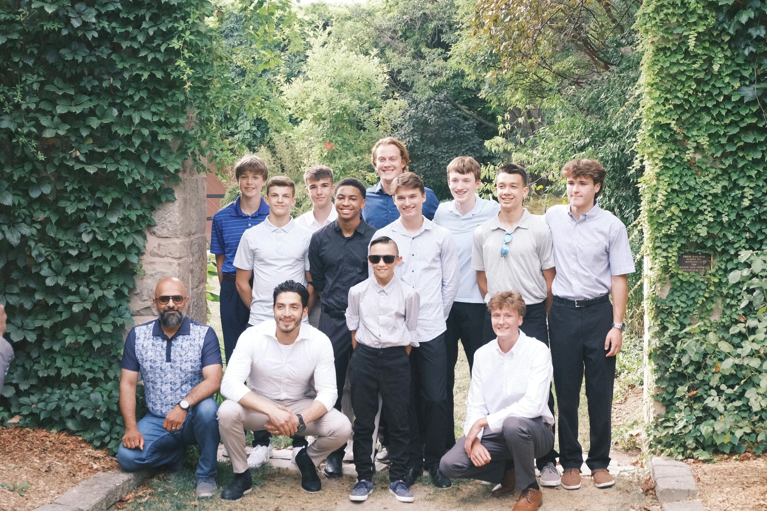 Group photo of young men and boys, some wearing sunglasses, standing and sitting outdoors in a garden with green foliage and stone pillars.