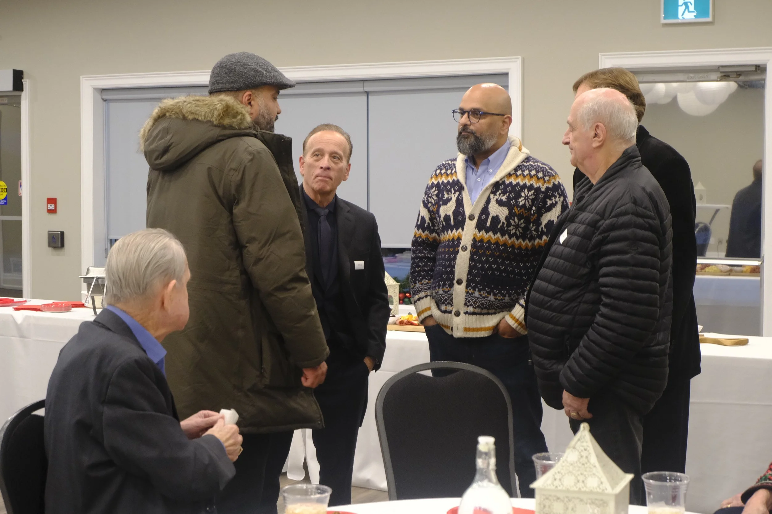 Group of men having a conversation at a holiday gathering in a indoor setting, with Christmas-themed sweater worn by one man, table with plates and drinks in the foreground, background with a table of food.