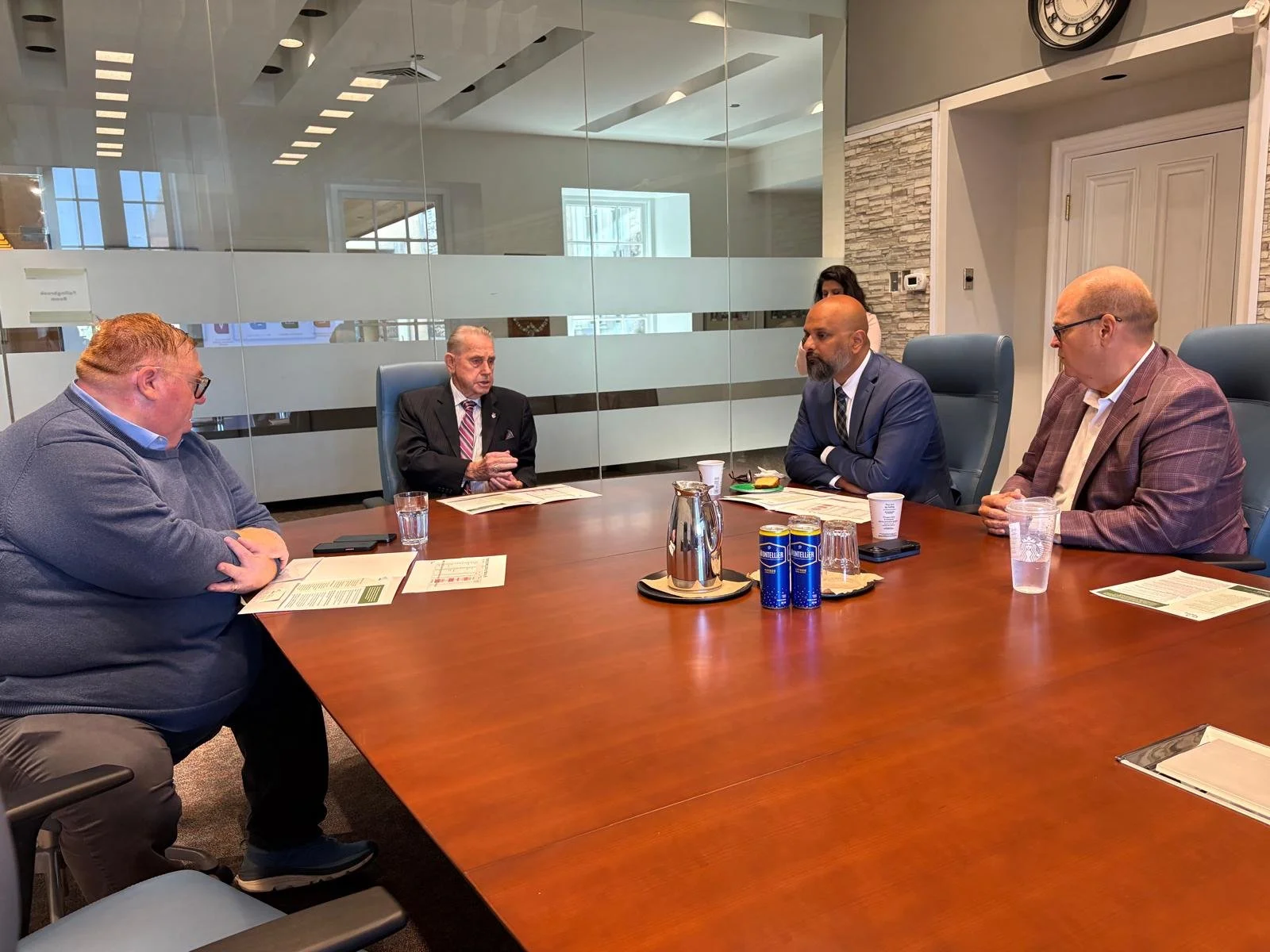 A business meeting with five men seated around a large conference table in a modern office. The man on the left is speaking, and the others are listening.