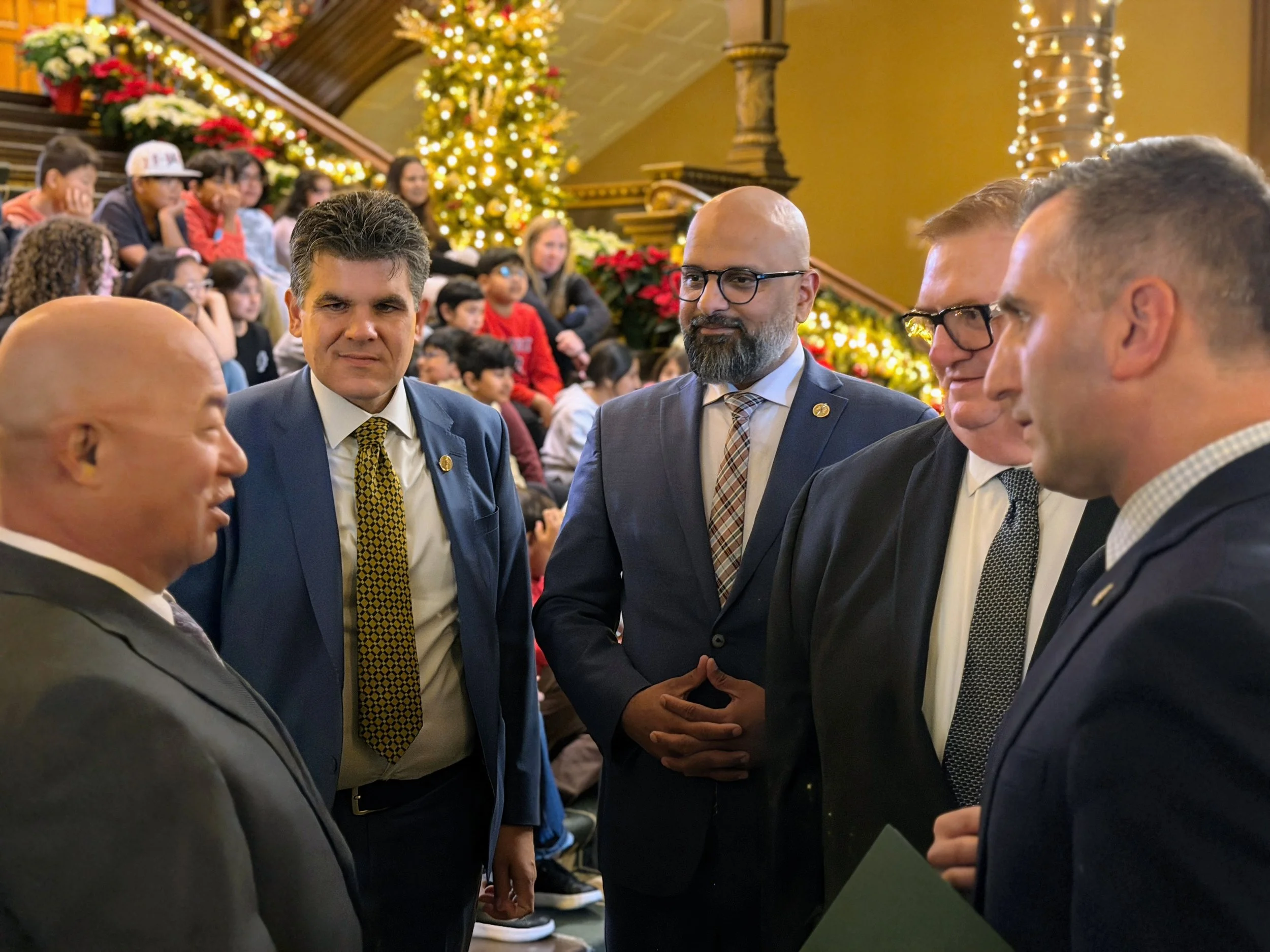 Five men in suits engaged in conversation at a festive indoor event with a Christmas tree and children in the background.
