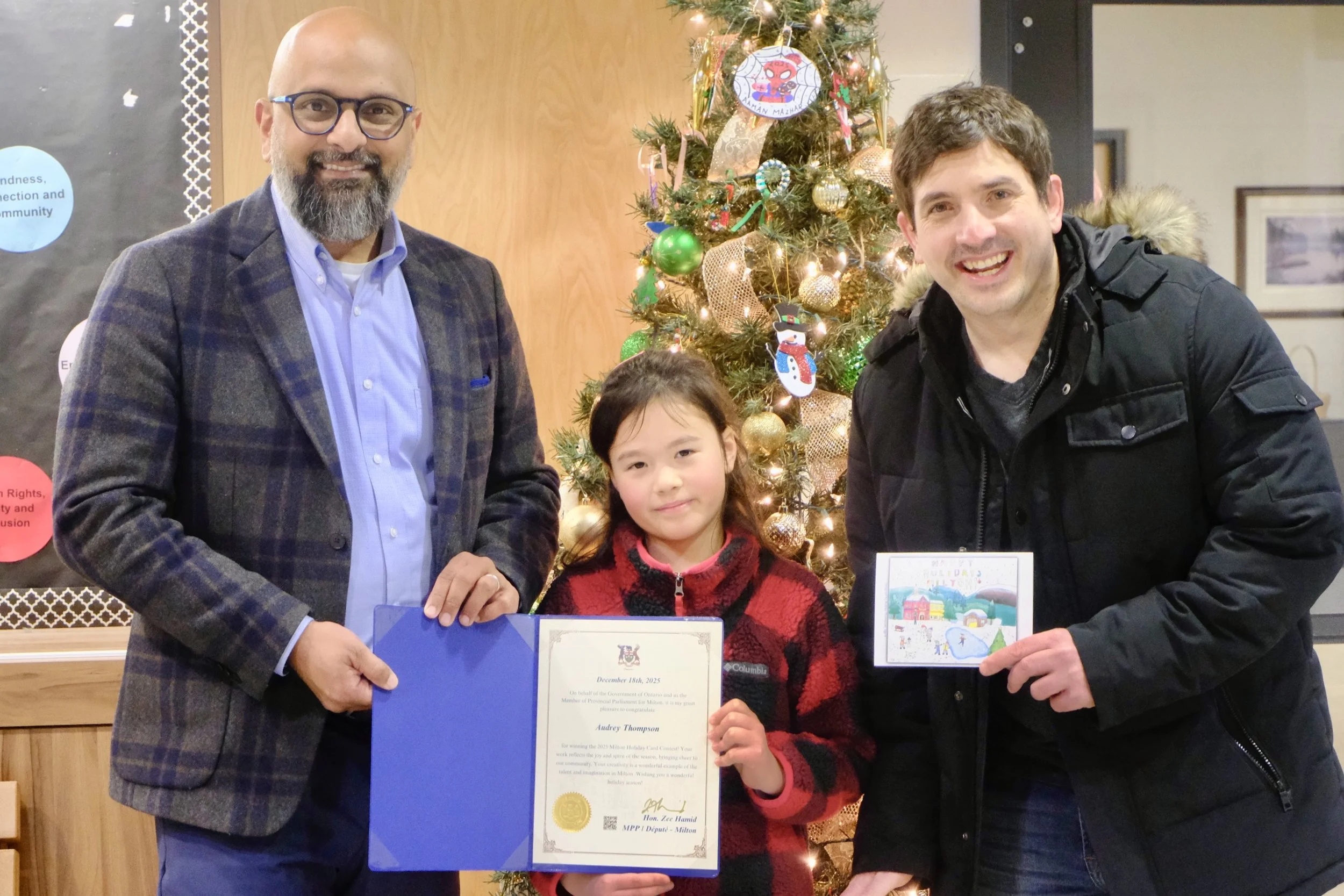 Three people, a man, a girl, and a man, stand in front of a decorated Christmas tree. The girl is holding a certificate, and the man on the right is holding a holiday card. All three are smiling.