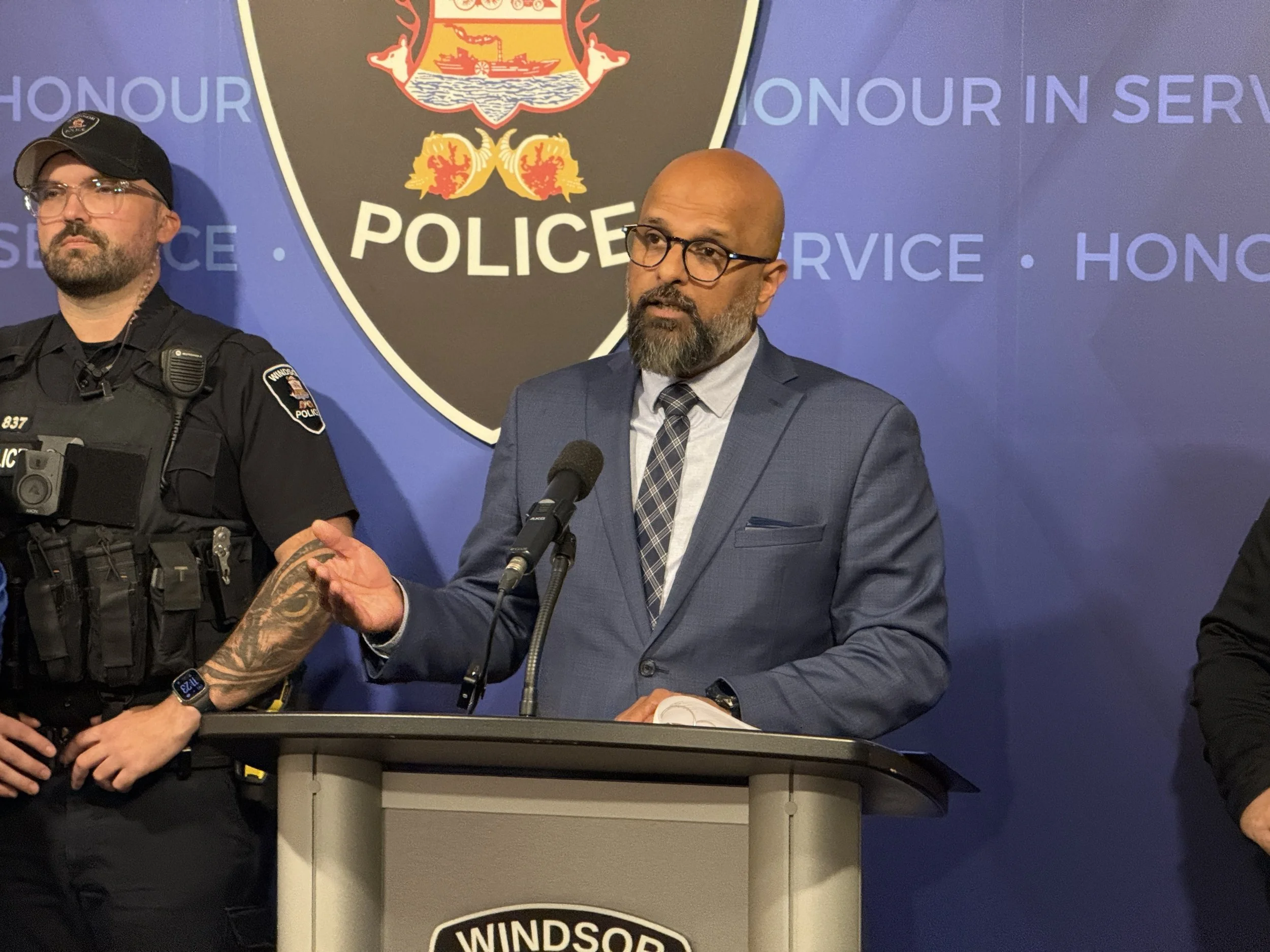 A man in a blue suit and glasses speaking at a podium with a police officer in uniform standing beside him. The backdrop displays a police emblem and the words 'POLICE' and 'HONOUR IN SERVICE'.
