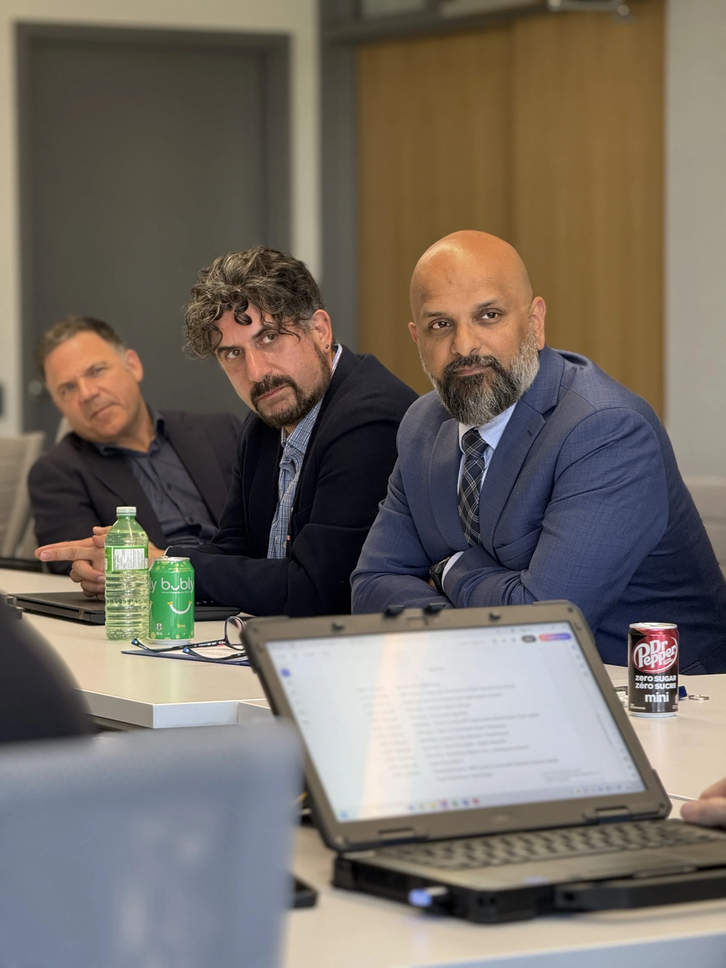 Three men in business attire seated at a conference table, engaged in a meeting, with laptops, drinks, and a tablet on the table.