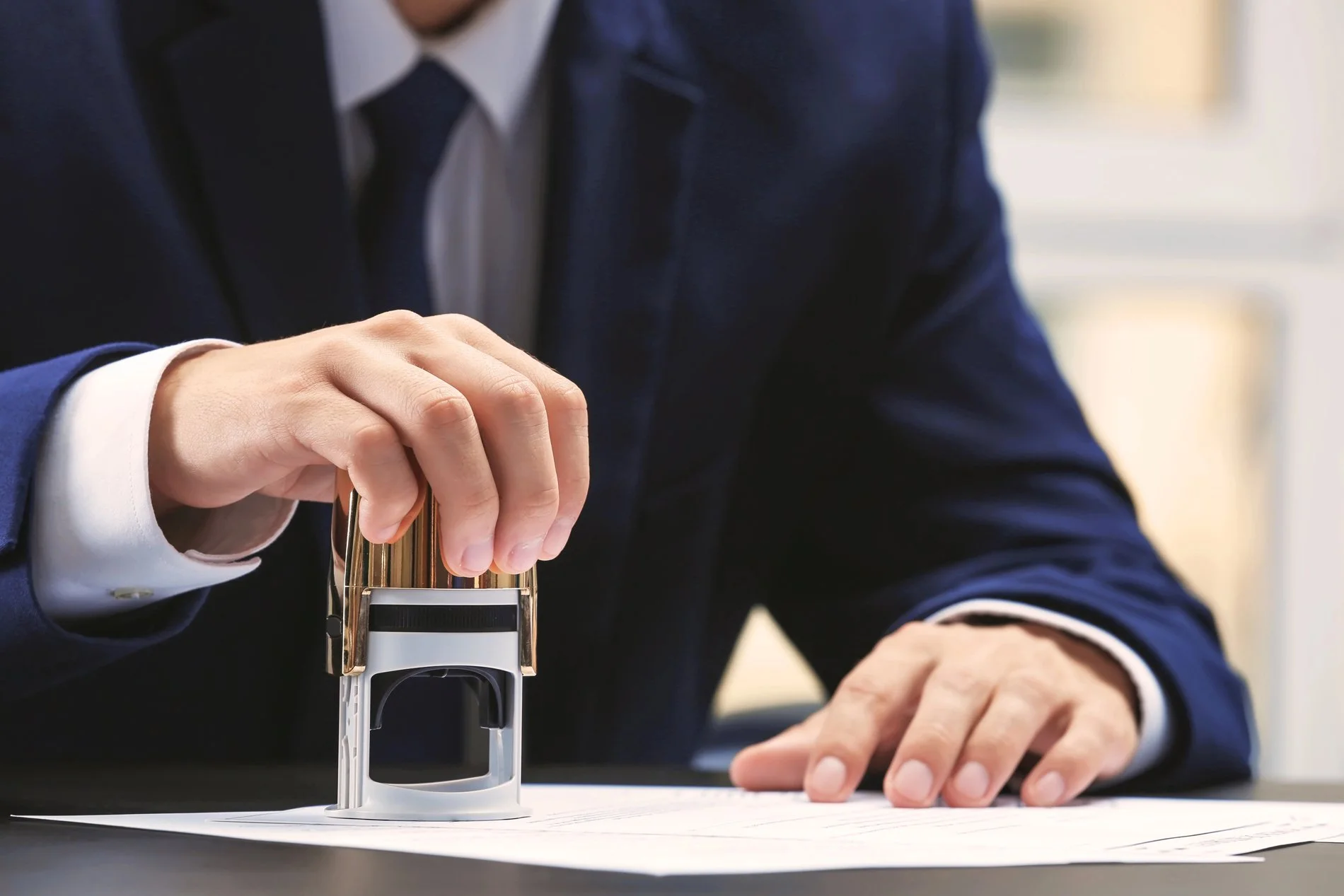 A person in a dark business suit using a red rubber stamp on a document.