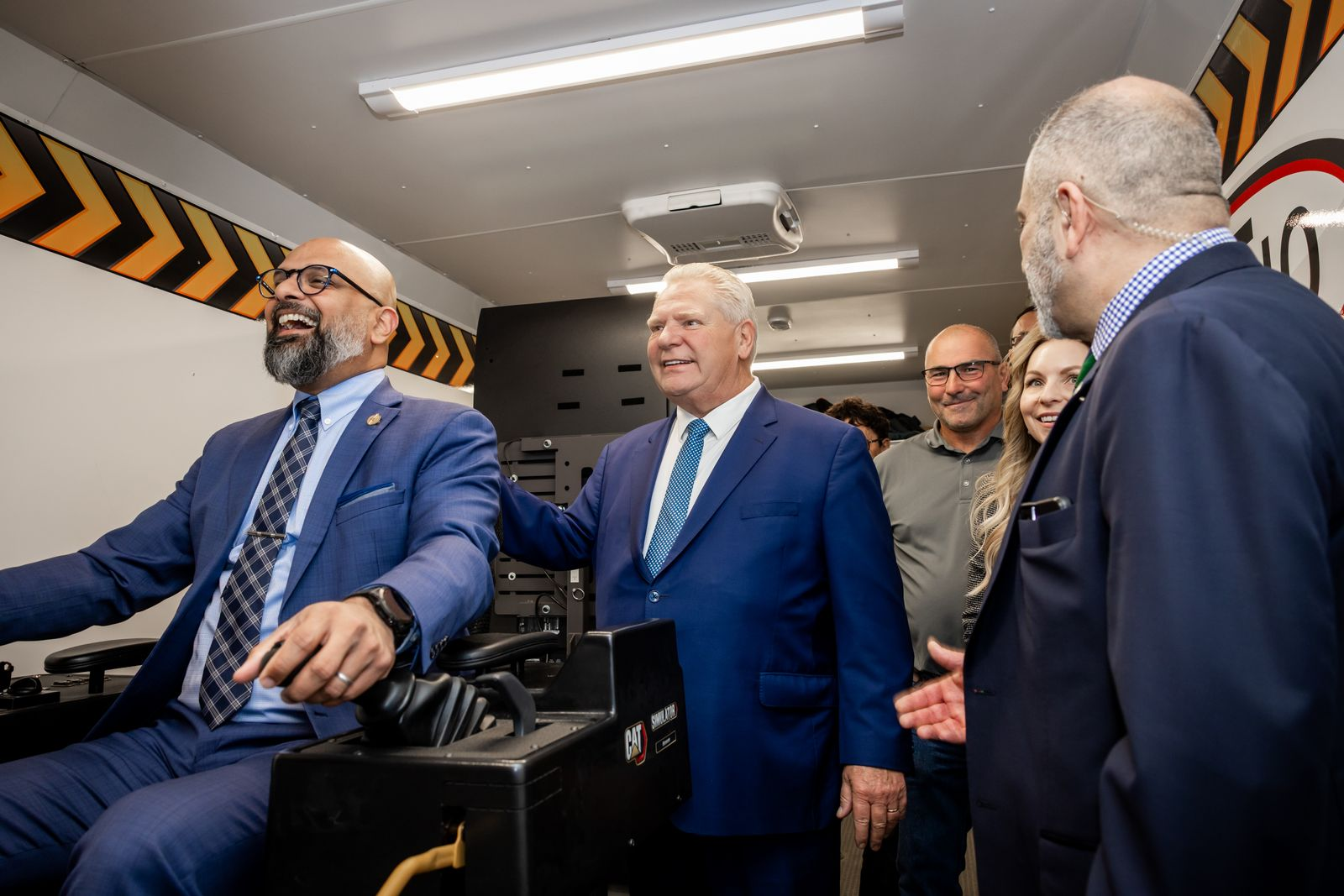 A group of professionally dressed people gathered in a room, smiling and engaging in conversation, with a focus on a man seated at a control panel or machinery.