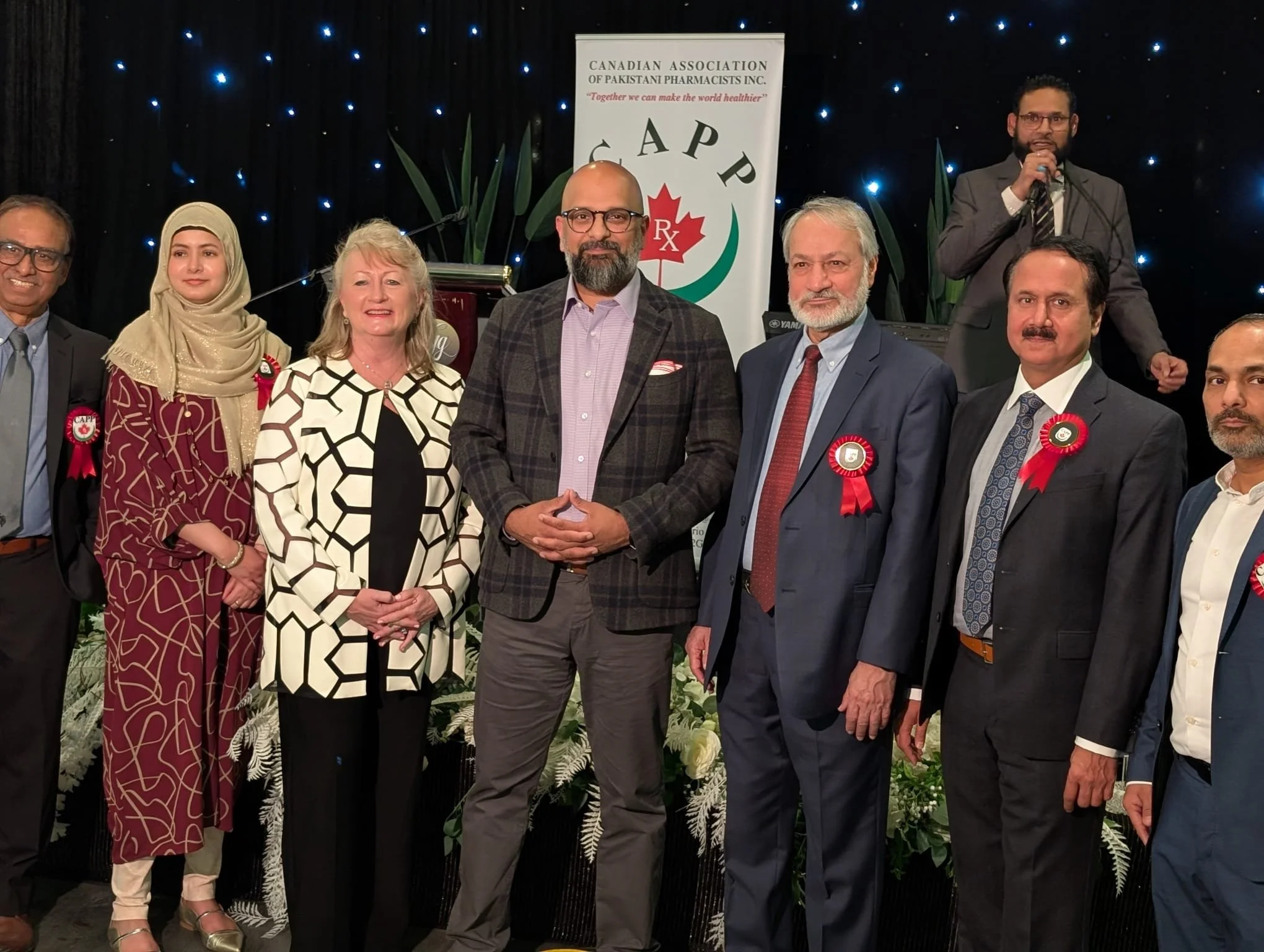 Group of nine diverse professionals on stage at a formal event, with a banner in the background that reads 'Canadian Association of Pakistani Pharmacists Inc.' and the slogan 'Together we can make the world healthier.' They are dressed in formal atti