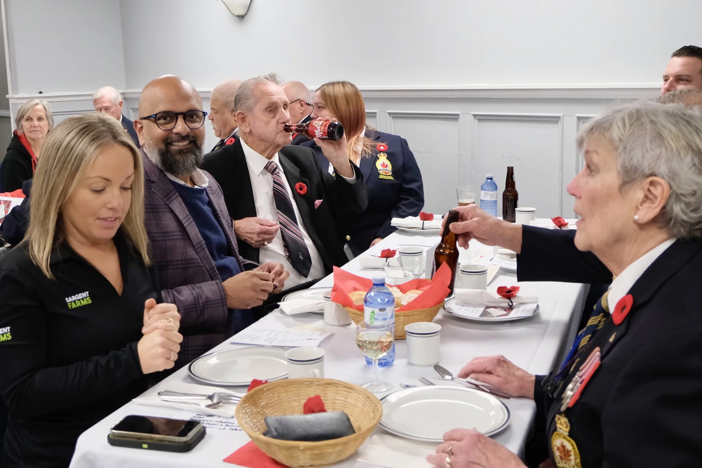 People sitting at a decorated dining table, celebrating, with some wearing poppy pins and military or veteran uniforms, one person drinking a soda, and various drinks and food items on the table.