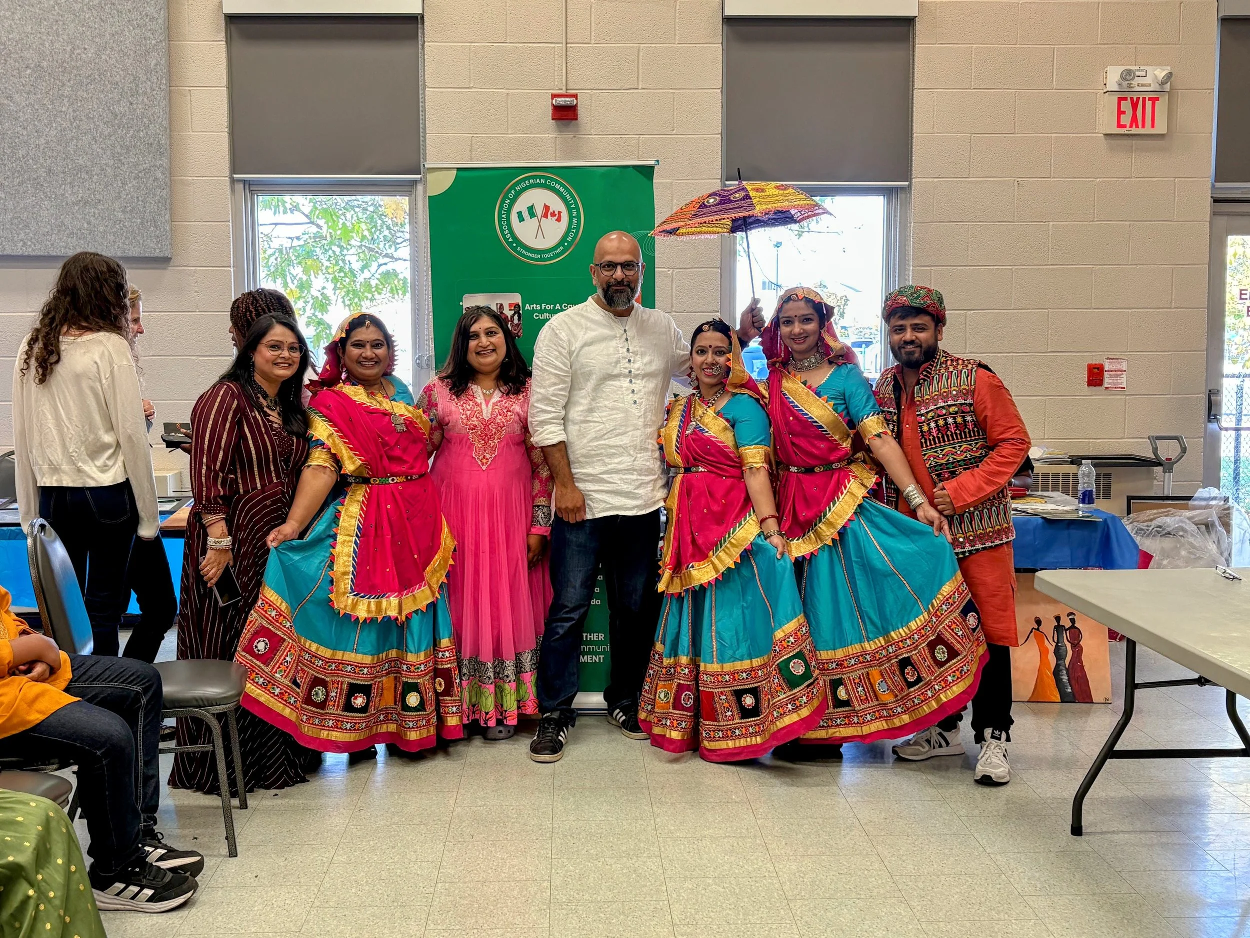 Group of people standing indoors, some in traditional colorful Irish dance costumes, with a man holding a colorful umbrella, posing for a photo at a cultural celebration event.