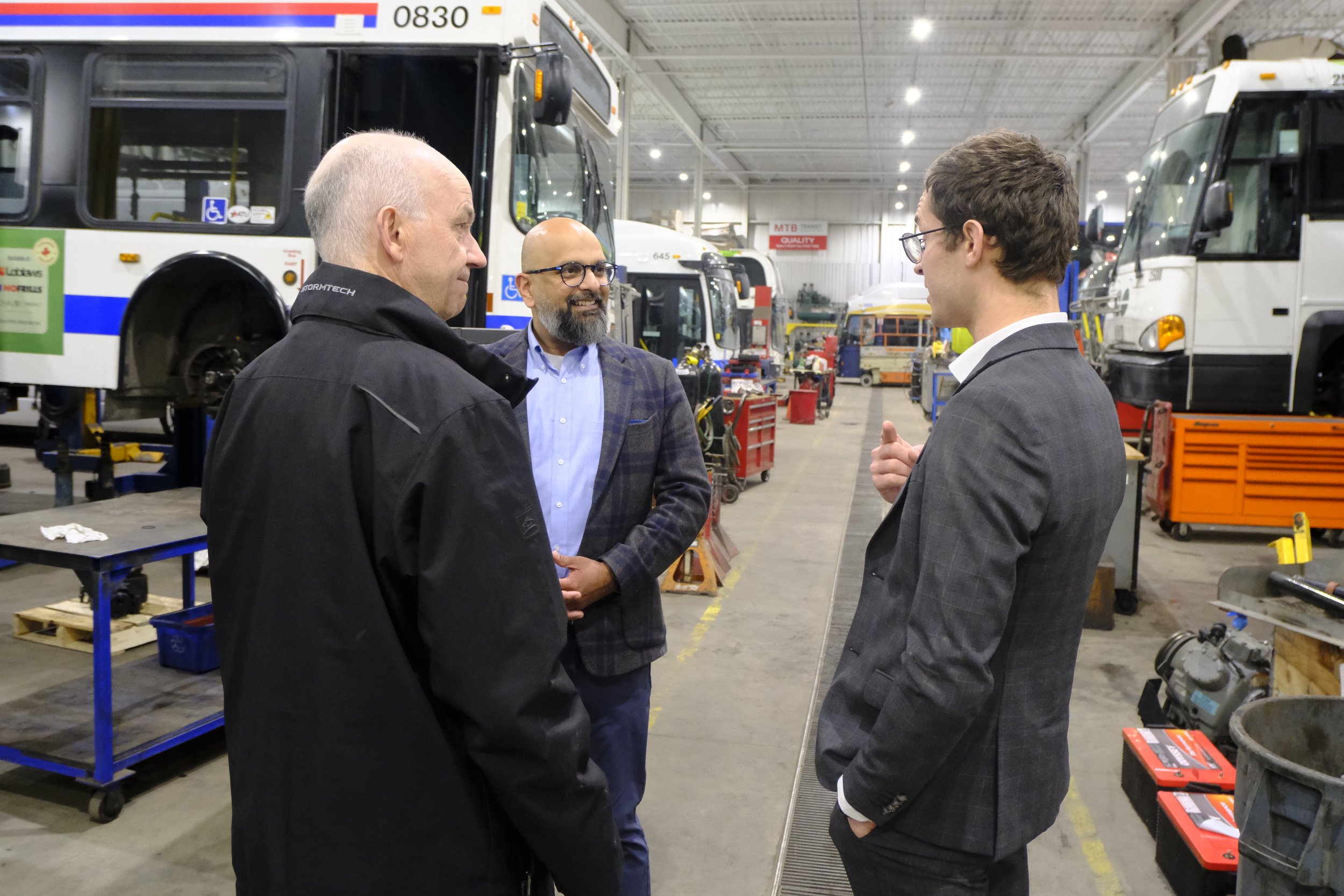 Three men in a garage or workshop engaged in conversation, with buses and various tools and equipment in the background.