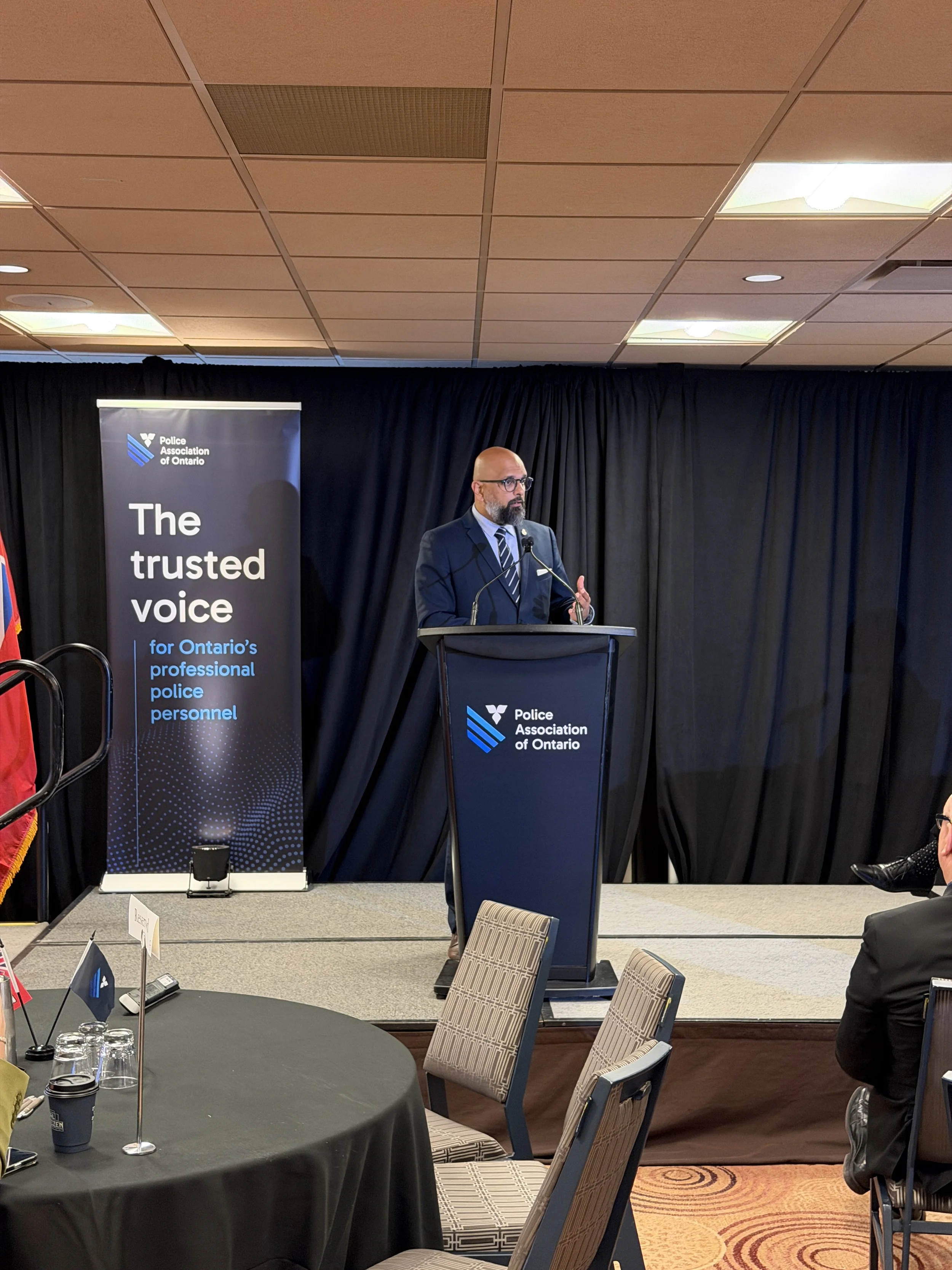 A man in a suit and glasses speaking at a podium during a conference, with a banner behind him that reads 'Police Association of Ontario,' and a black curtain backdrop.