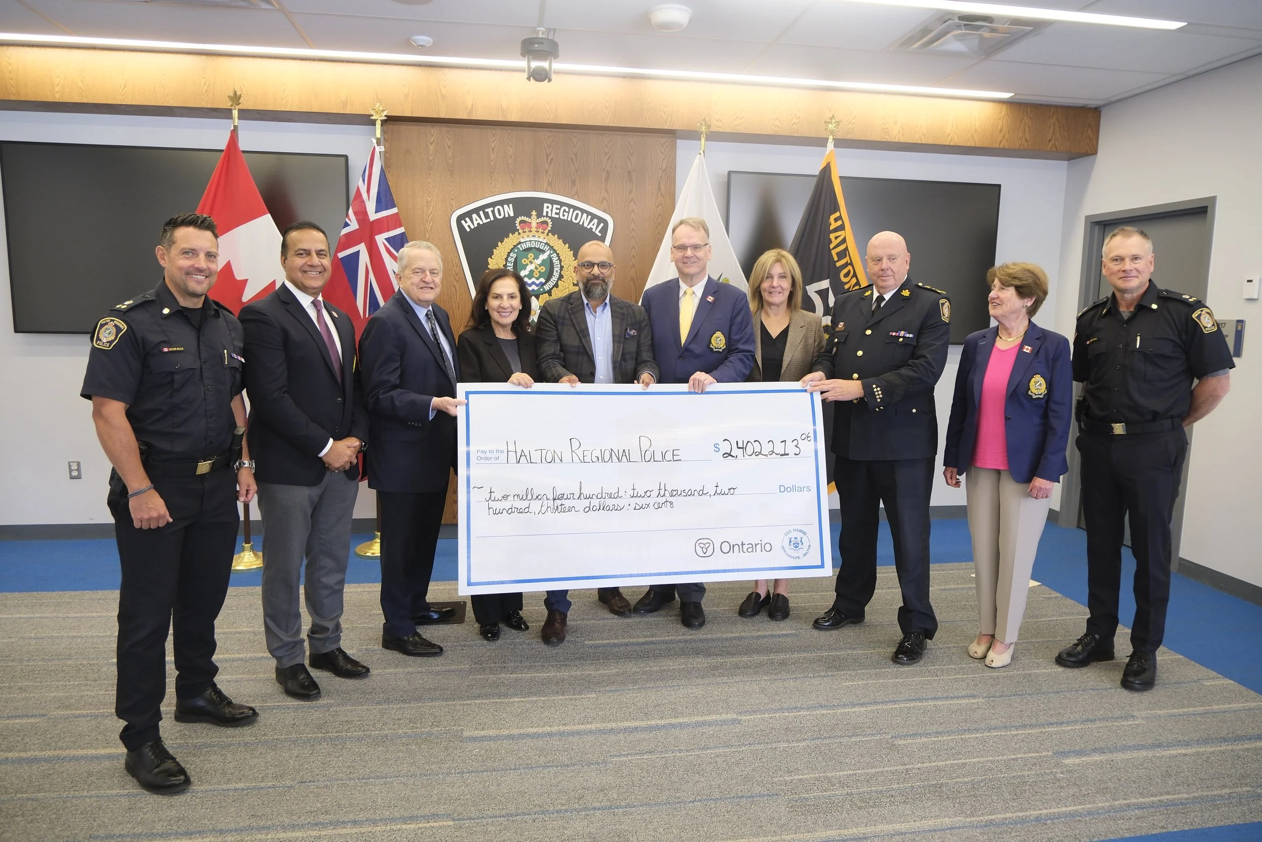 Group of police officers and officials holding a large ceremonial check for $2,402,213 at a police station, with flags and a police badge in the background.