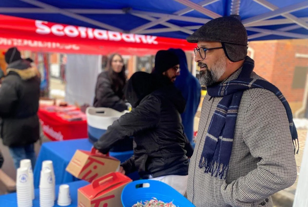 A man with glasses and a beard wearing a coat, scarf, and flat cap standing at an outdoor booth with a red and blue canopy, talking to other people during an outdoor event.
