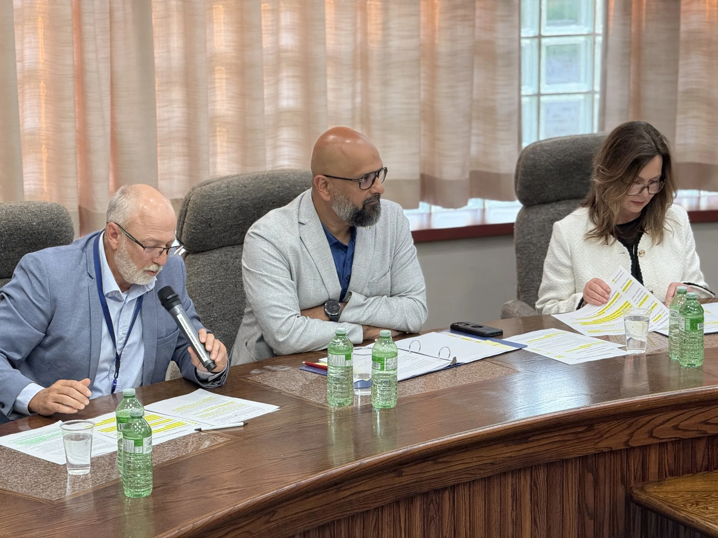 Three people sitting at a conference table with documents, water bottles, and glasses. The person on the left, an elderly man with a white beard and glasses, holds a microphone. The middle person, a man with glasses and a beard, looks ahead. The pers
