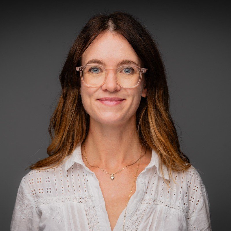 A woman with shoulder-length brown hair and glasses, wearing a white embroidered blouse and a delicate necklace, smiling against a dark grey background.