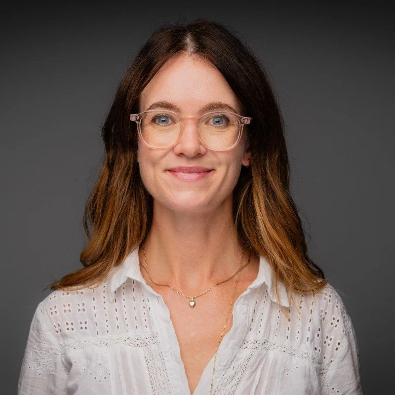 A woman with shoulder-length brown hair wearing glasses and a white blouse, smiling against a dark gray background.
