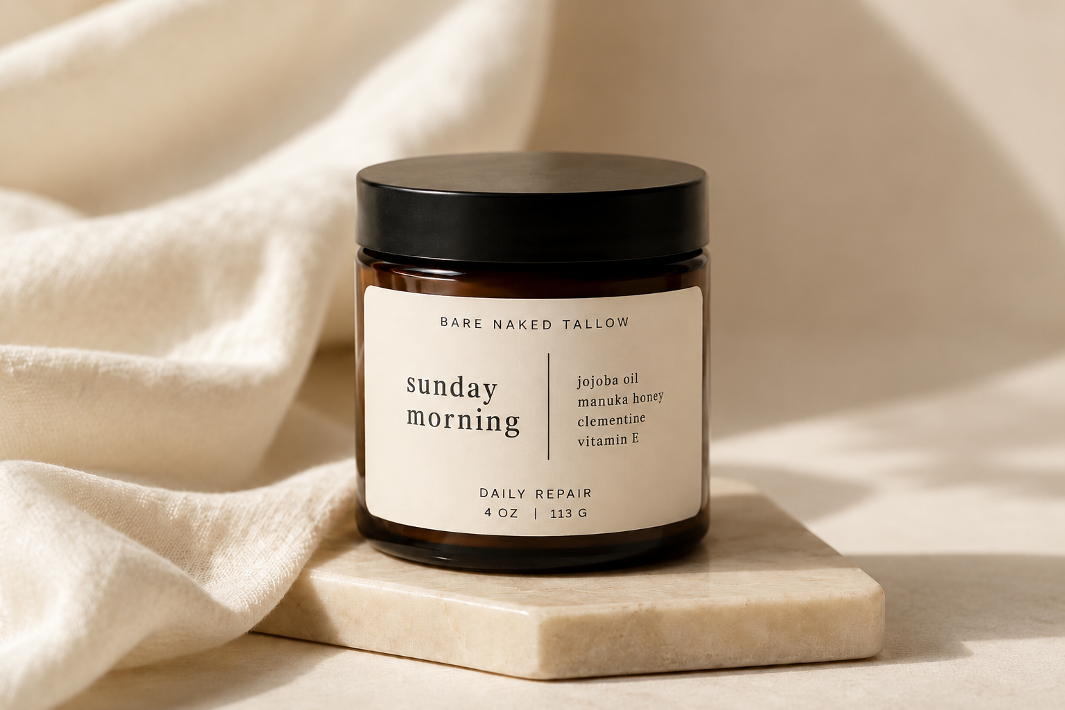 A brown jar with a black lid, labeled 'Sunday Mourning' with ingredients listed as premium tallow, Manuka honey, and clementine, resting on white satin fabric