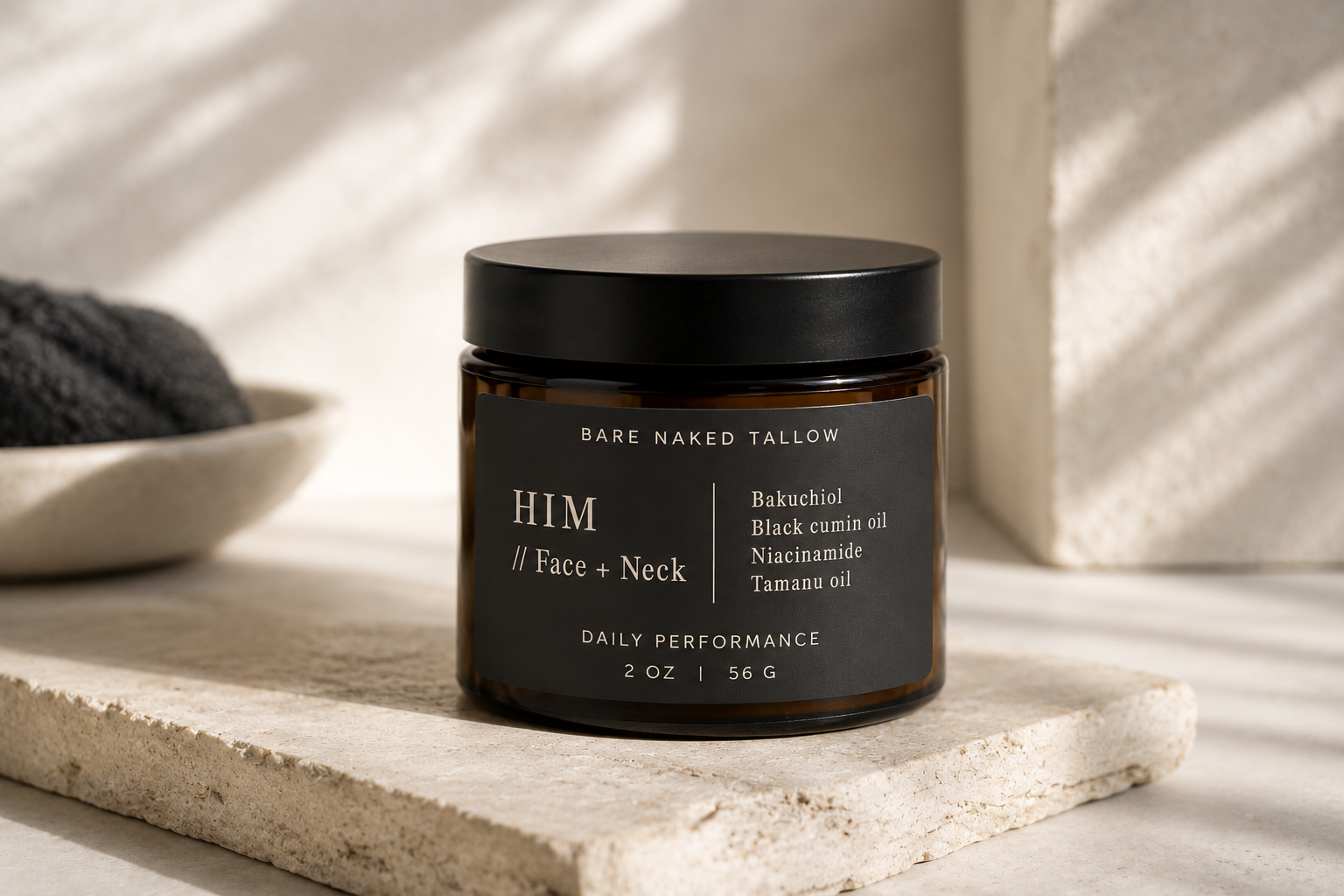 A dark brown jar with a black lid labeled 'HIM FACE & NECK with Sea Grass, Aloe & Neroli' sitting on a textured stone surface, surrounded by black fabric and green leaves.