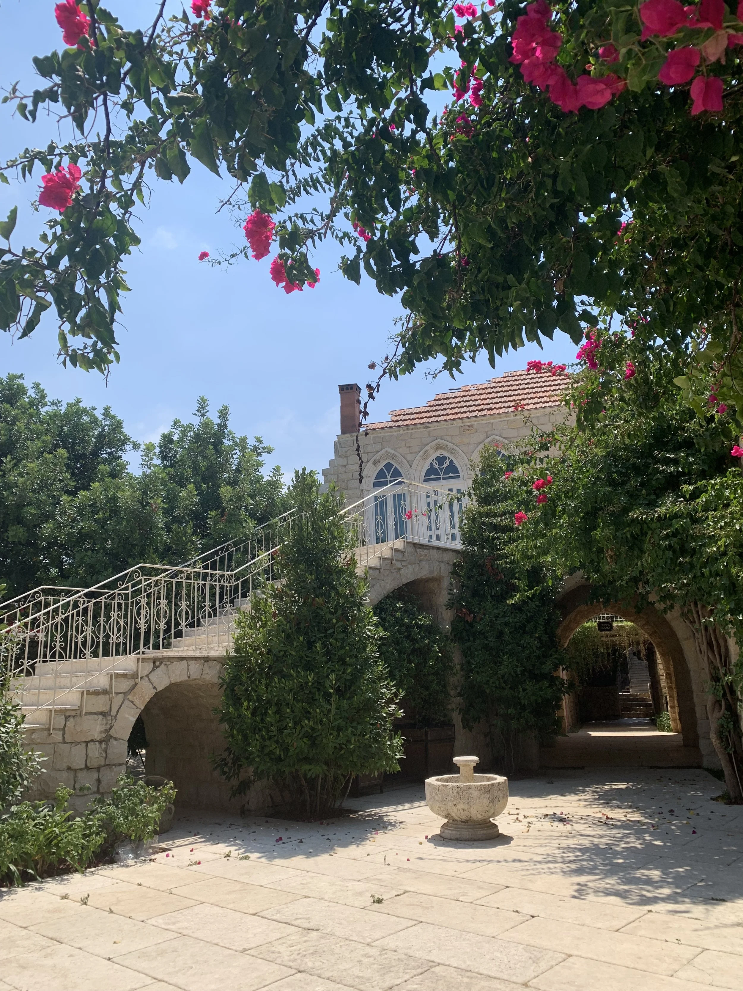 A white-stone house with traditional pointed Lebanese windows sits atop a tall stone staircase and is surrounded by trees and dangling flowers.