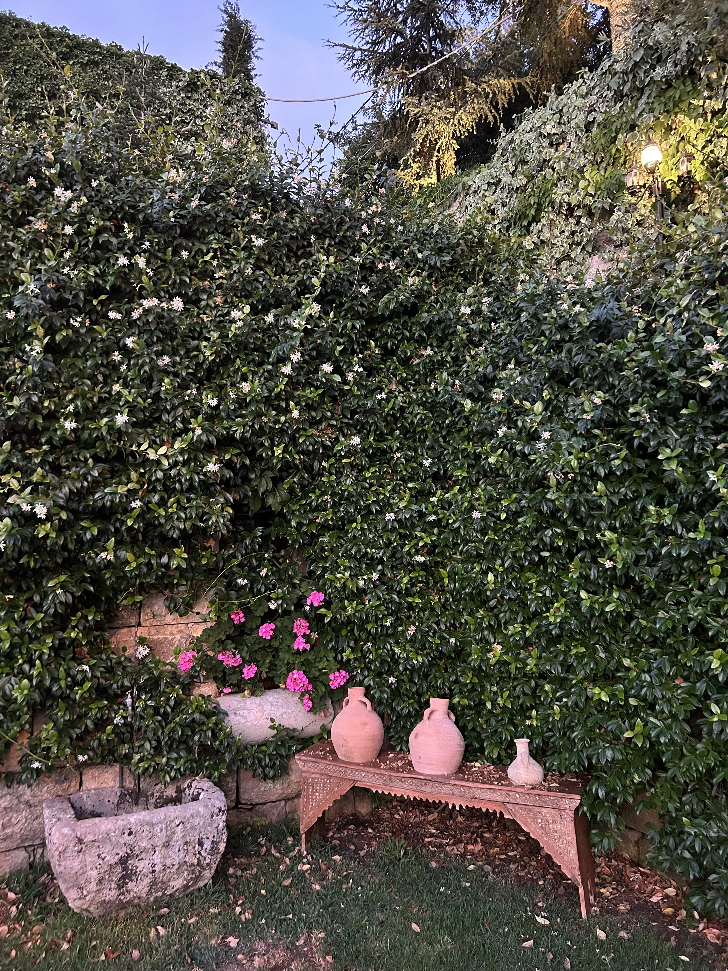 A traditional Lebanese wood-inlaid bench sits under three clay pots. The scene is surrounded by blooming jasmine vines.
