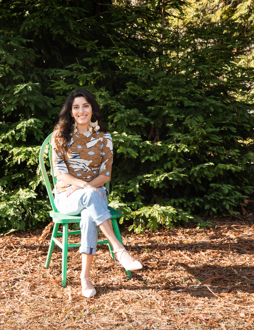 Tania sits with her legs crossed in a green chair in front of a large Redwood Tree. She has a gentle smile and is wearing a flowery top and jeans.