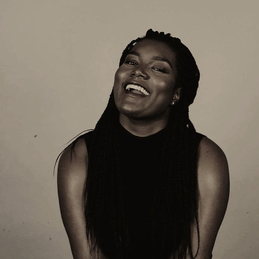 A woman with dark skin and long braided hair smiling and laughing against a plain neutral background.