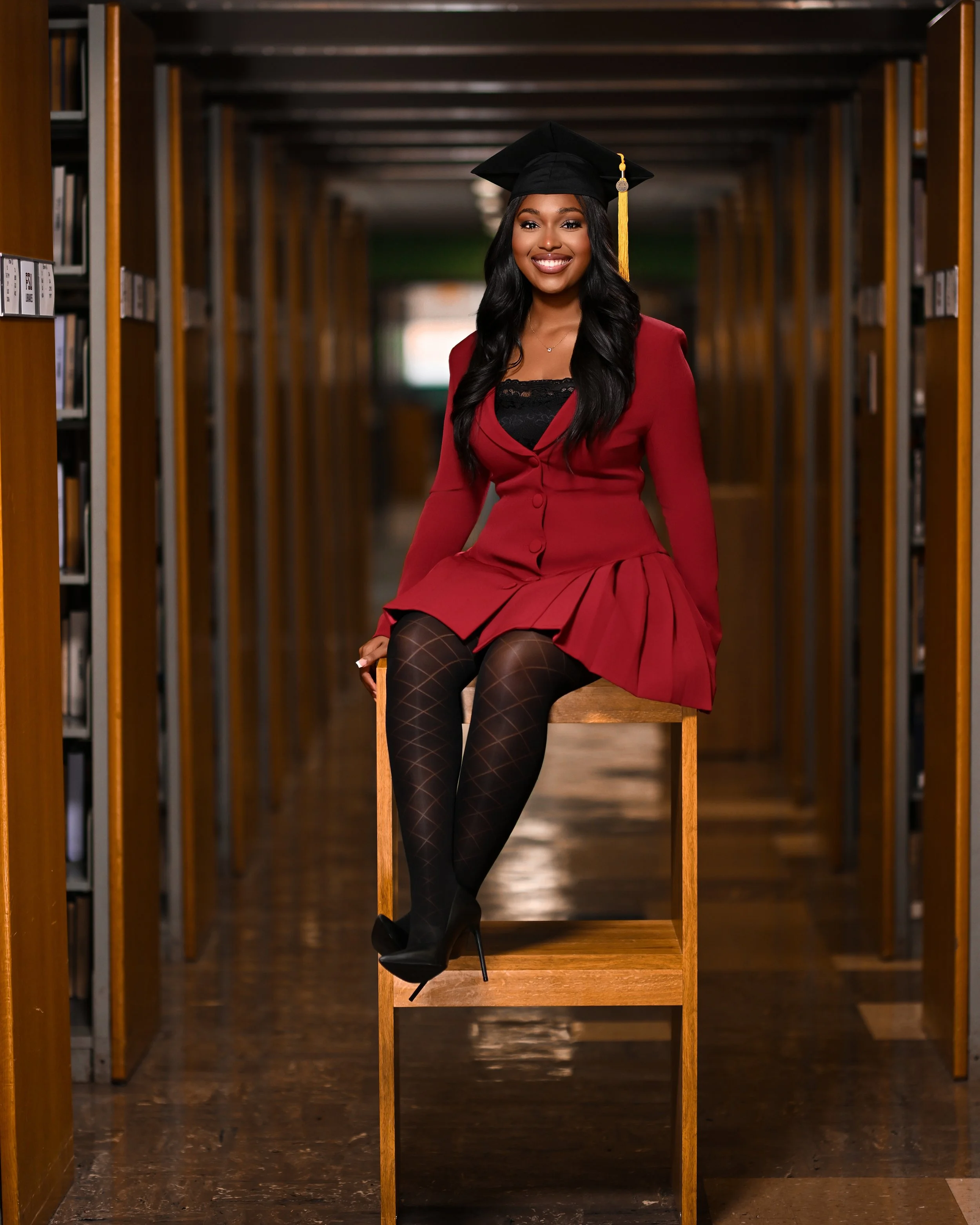 Smiling young woman in a graduation cap and red dress sitting on a wooden chair in a library aisle, with bookshelves on either side.