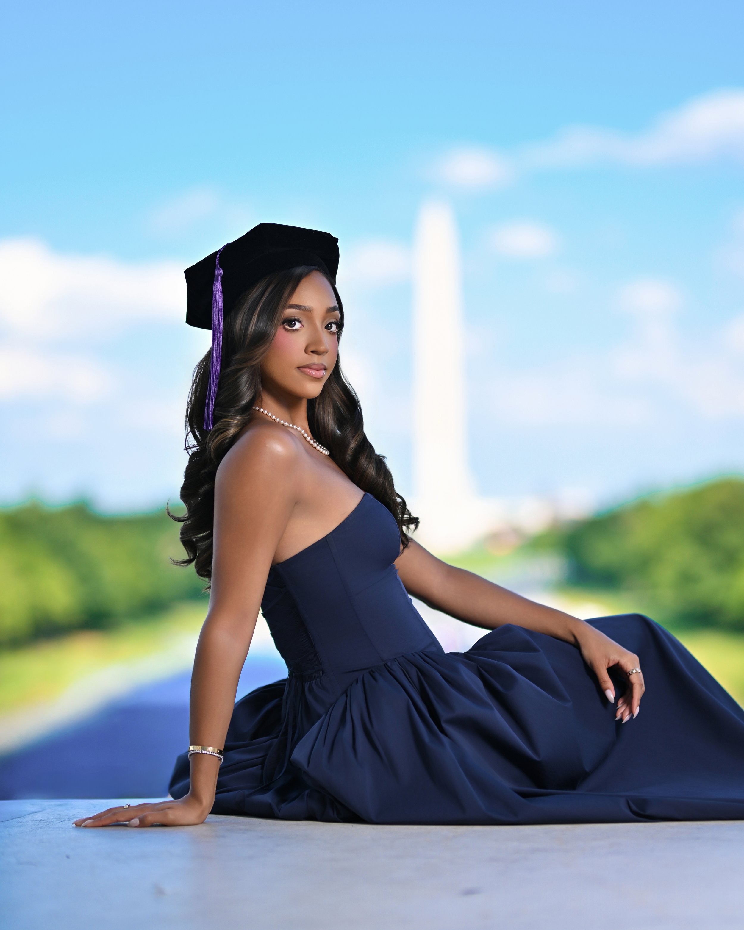 A young woman in a navy blue strapless dress and a graduation cap with a purple tassel, sitting on the ground in a graduation photo shoot outdoors with a blurred background of a blue sky and green landscape.