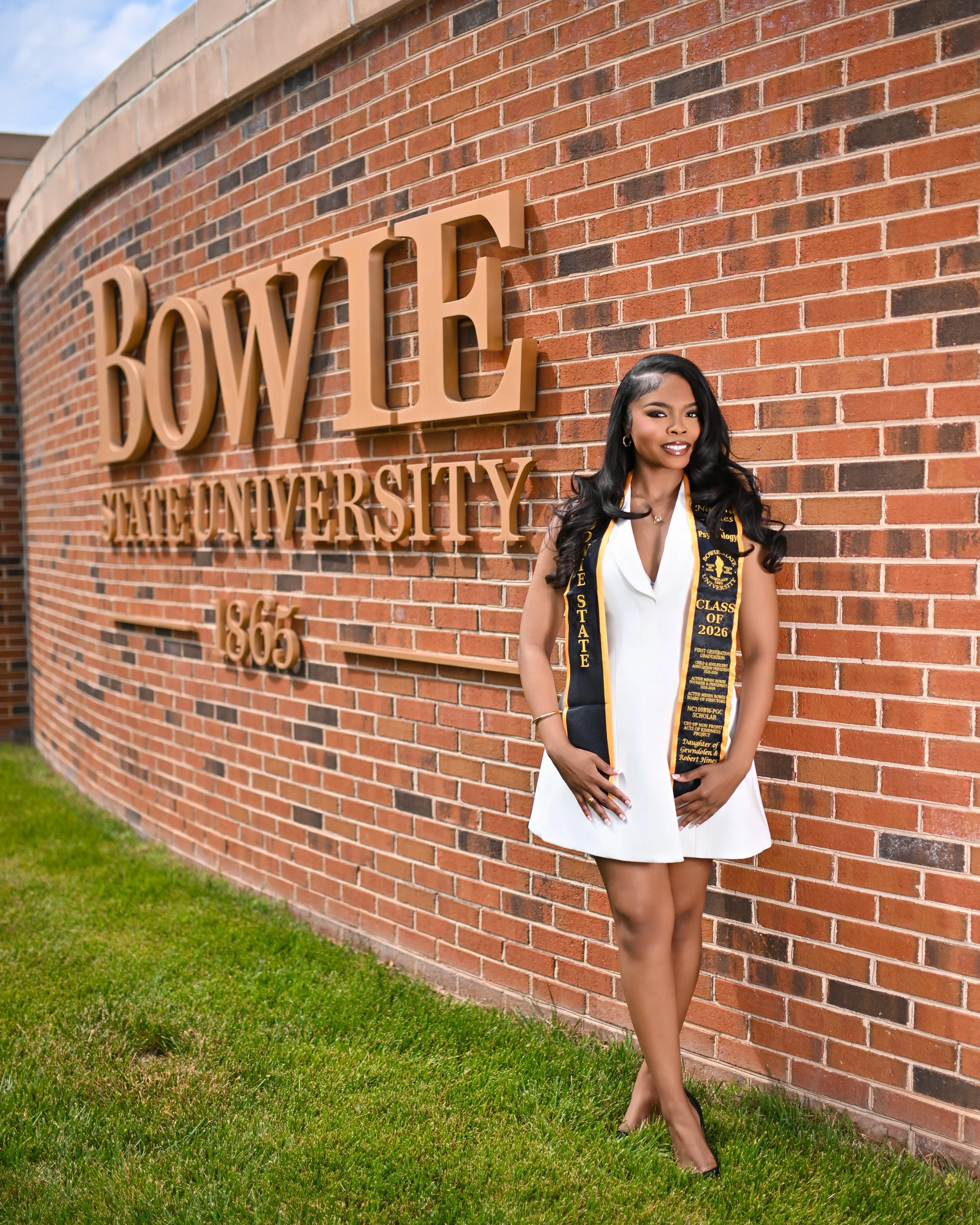 A young woman with long black hair, dressed in a white dress and black high heels, smiling and standing against a red brick university wall, wearing a black and gold graduation stole and sash that read 'Class of 2026'.