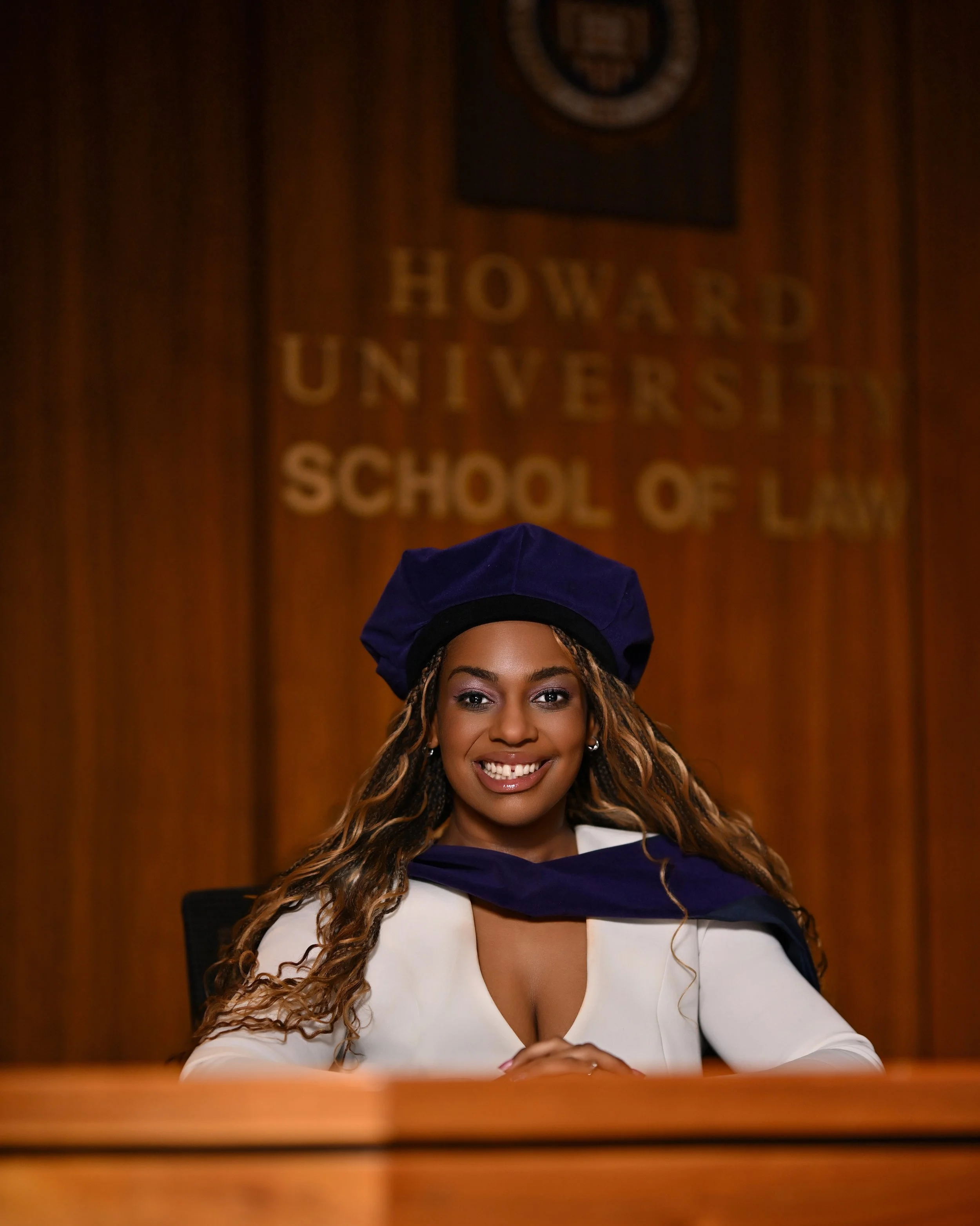 Woman in graduation cap and gown sitting at a table in a wood-paneled room, with the Harvard University School of Law emblem on the wall behind her.