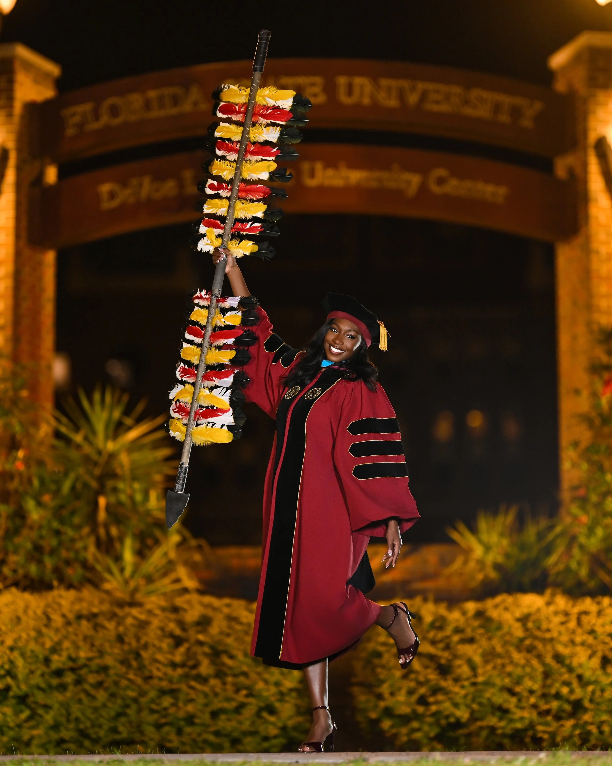 Female graduate in maroon cap and gown holding a colorful feathered staff at a graduation ceremony at the University of Florida, nighttime.