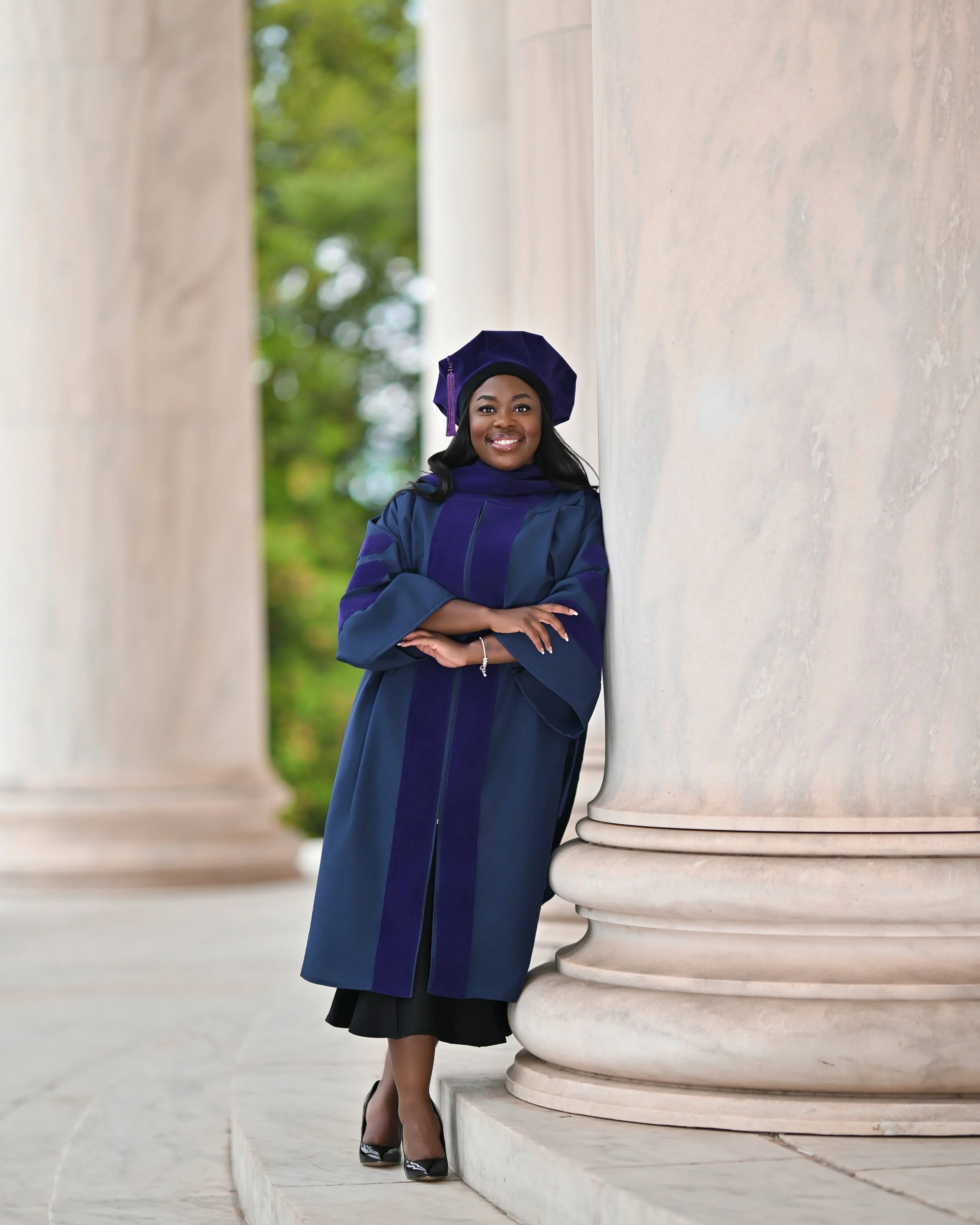A woman wearing a graduation gown and cap standing outside next to a large stone column, with greenery in the background, smiling and posing with her arms crossed.